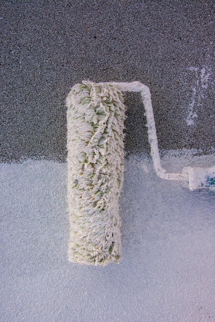 Worker applies acrylic white paint on the roof of the house. Hand of dark skinned worker holding a roller applies white coating paint to insulate the roof of his house.