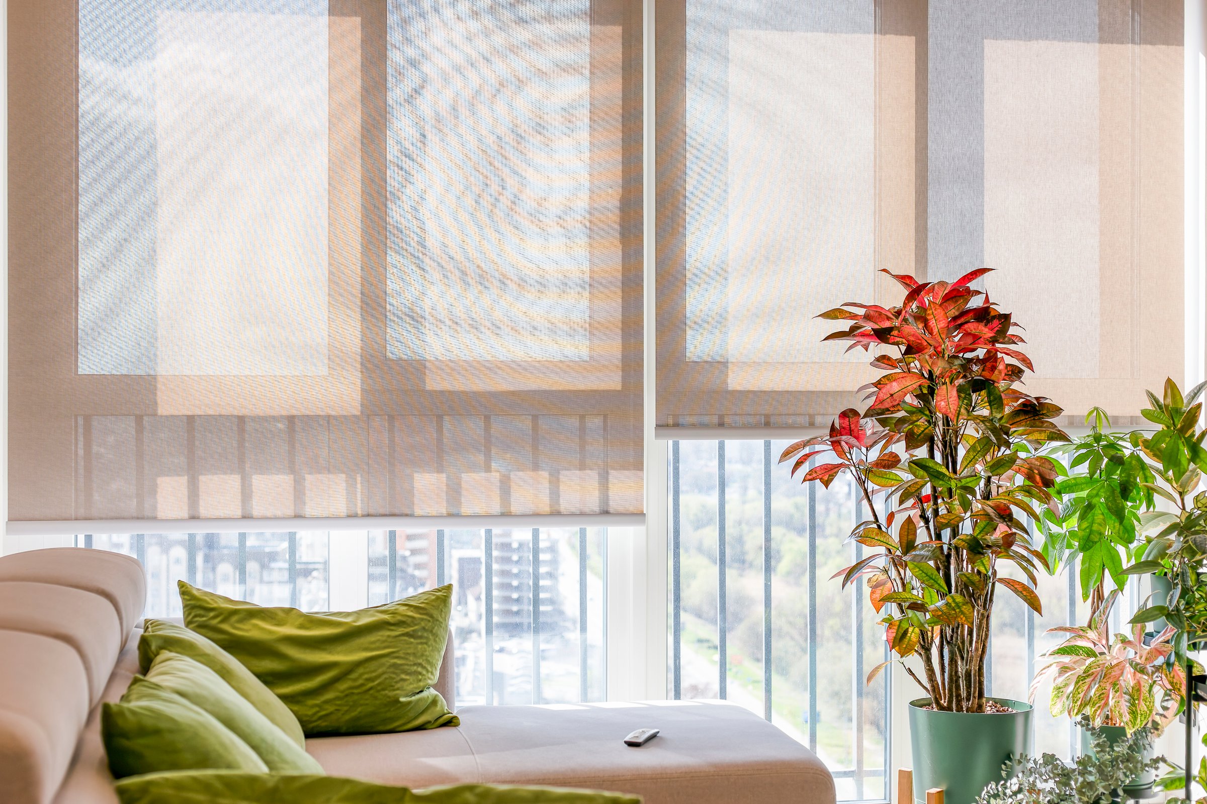 Automatic roller shades with screen material cover large windows, filtering sunlight into a living space. Motorized sunscreen blinds beige color with remote control panel on a sofa. Cityscape outside.