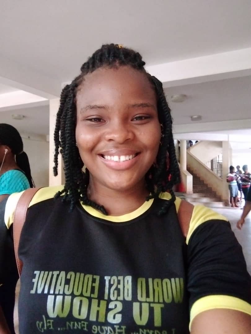 Smiling woman with braided hair wearing a black T-shirt with yellow trim at an indoor location.