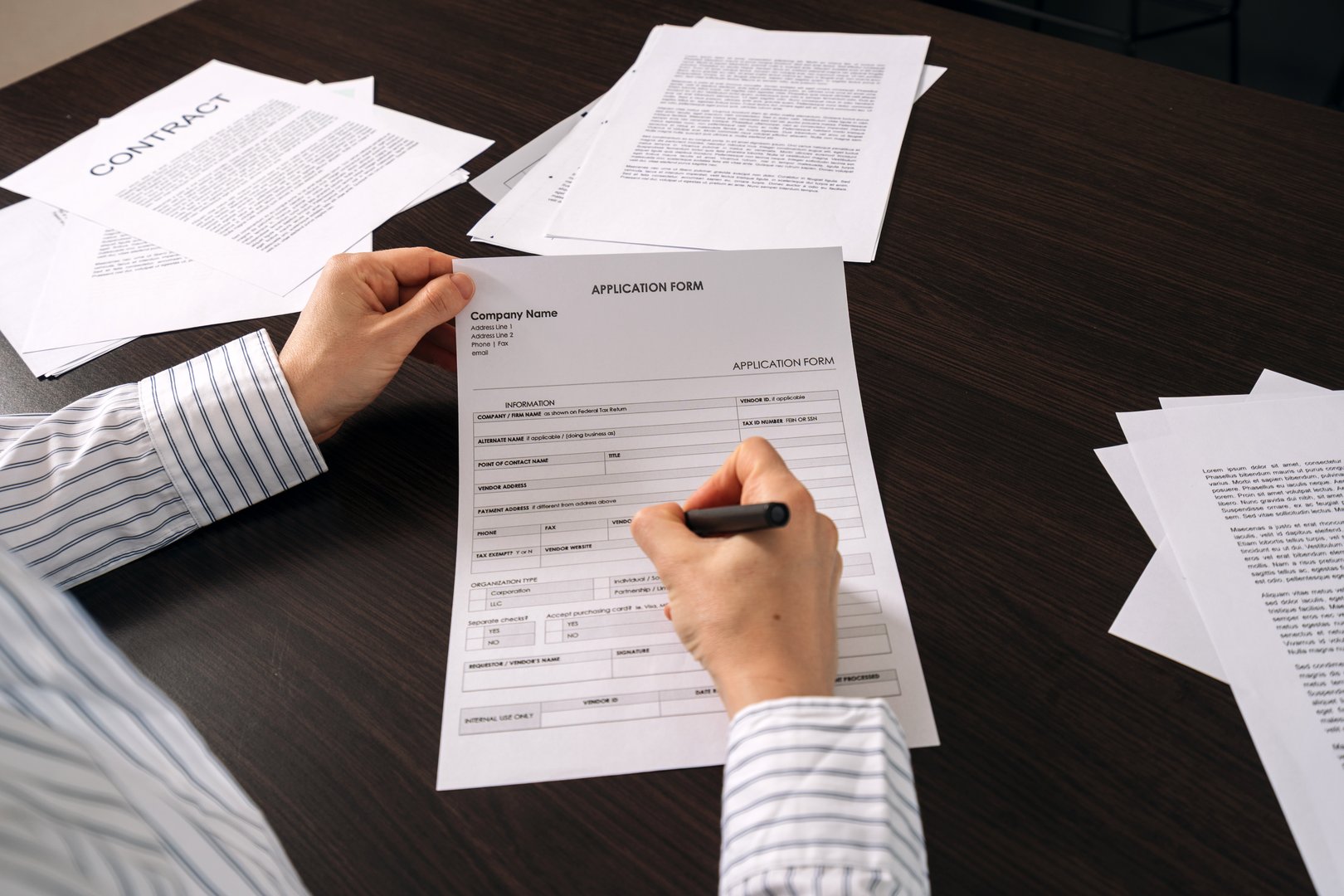 Student apply for college or university. POV, a person is completing an application form with a pen on a desk. Several documents, including contracts and reports, are scattered around the workspace