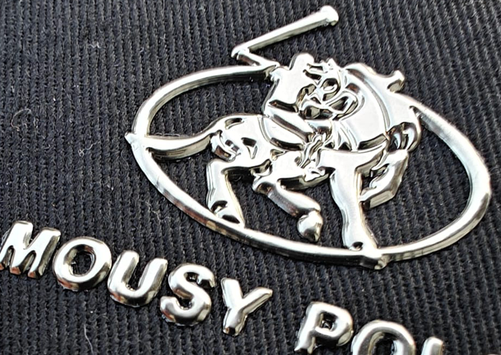 Close-up of a silver polo player emblem with the word MOUSY POL on a textured black fabric background.