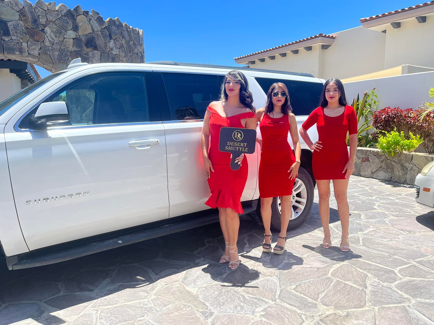 Three women in red dresses stand by a white SUV, holding a Desert Shuttle sign in sunny outdoor setting.