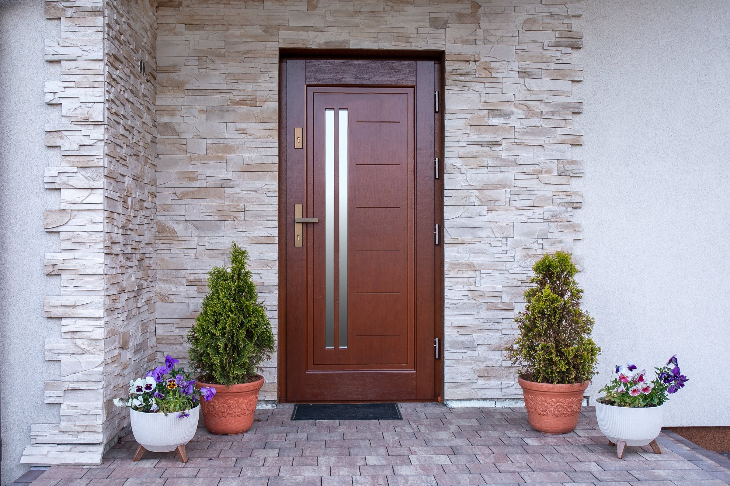 A front door detail with a stone accent wall, a brown door, covered porch, white posts and railing, and white siding. Exterior facade. External doors. Wooden doors.