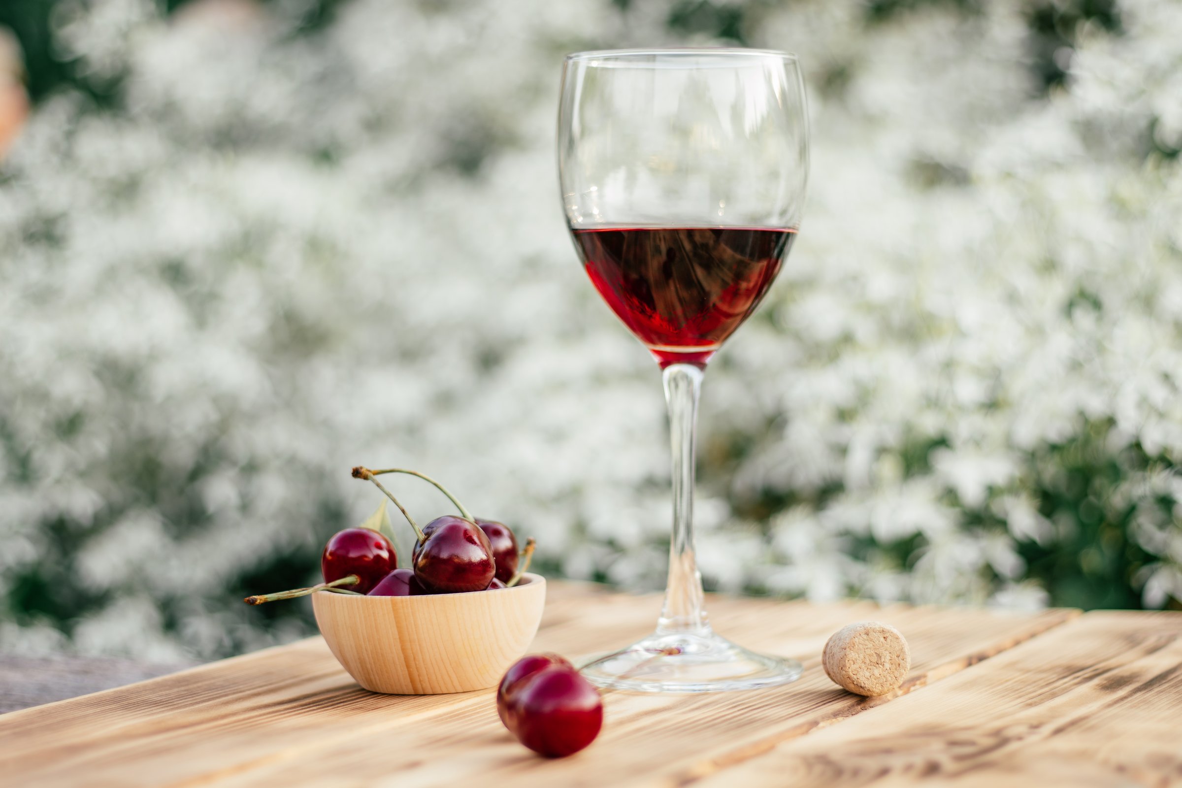 half-empty glass of red wine, wine stopper and sweet cherries in wooden cup on table in sunny day in garden against white organic background. selective focus