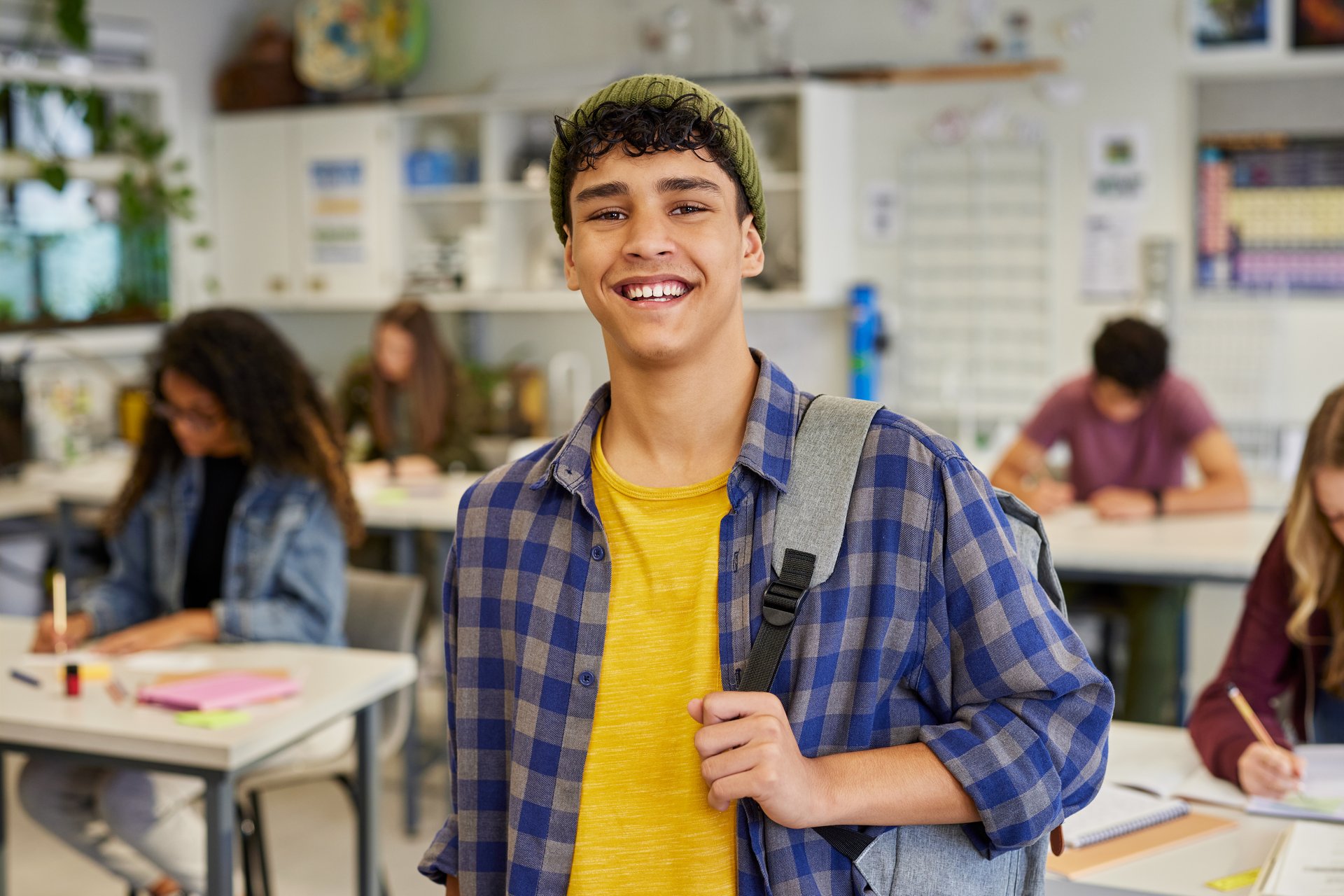 Happy male student standing in classroom with backpack and casual outfit while looking at camera. Confident multiethnic guy smiling while classmates study in background at secondary school. Successful mixed race teenager standing with backpack and wearing green beanie with a big smile.