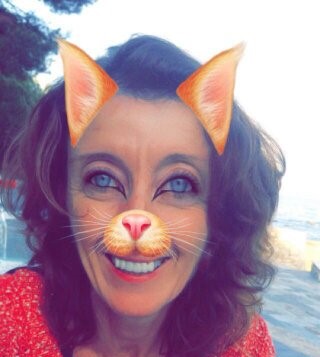 Person with a cat face filter, featuring cat ears and nose, smiling outdoors with a background of trees and sky.