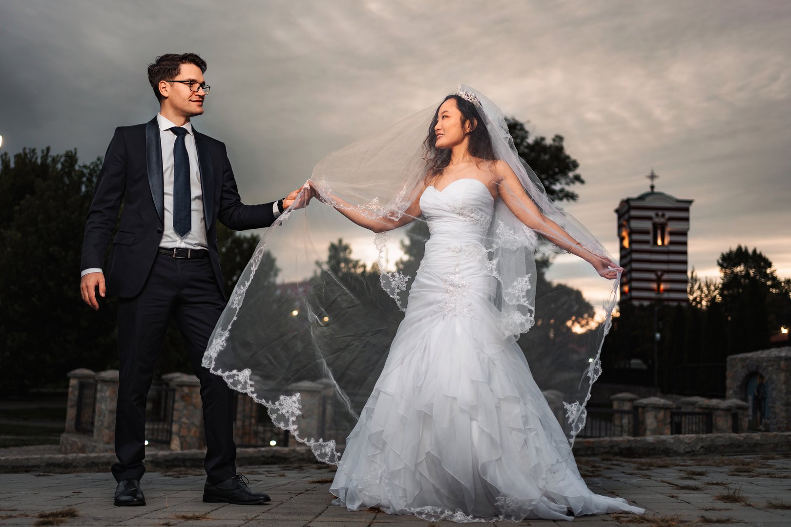 Groom holding bride's flowing veil outdoors at dusk, celebrating their wedding day