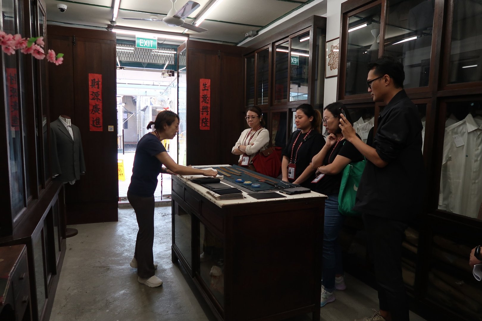 A tailor showing fabric samples to a group of people in a clothing shop with suits and shirts displayed.