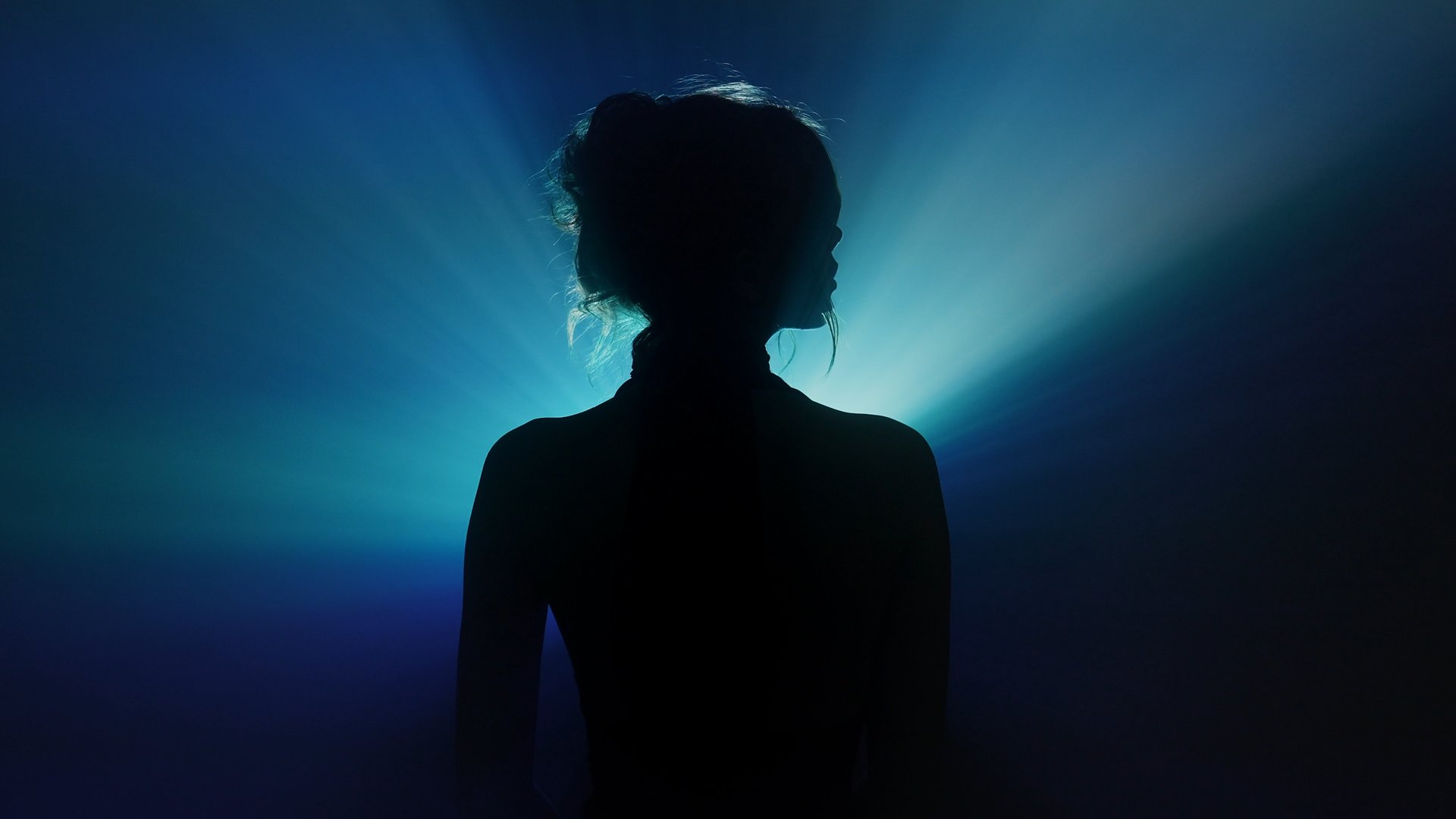 Mysterious silhouette of a woman standing against a dark background, surrounded by dramatic blue and teal light rays forming a radiant, cinematic halo.