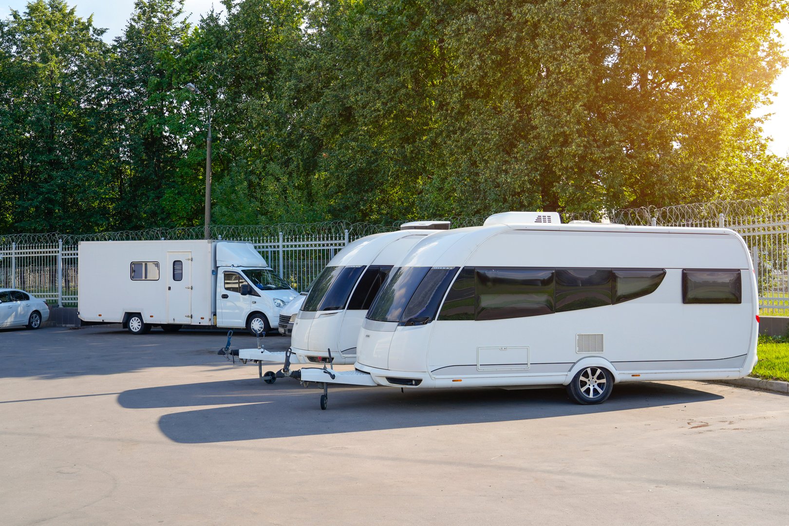 Two modern caravans are parked side by side in a sunlit area. Lush green trees surround the lot, creating a peaceful outdoor ambiance. A camper van is also visible in the background