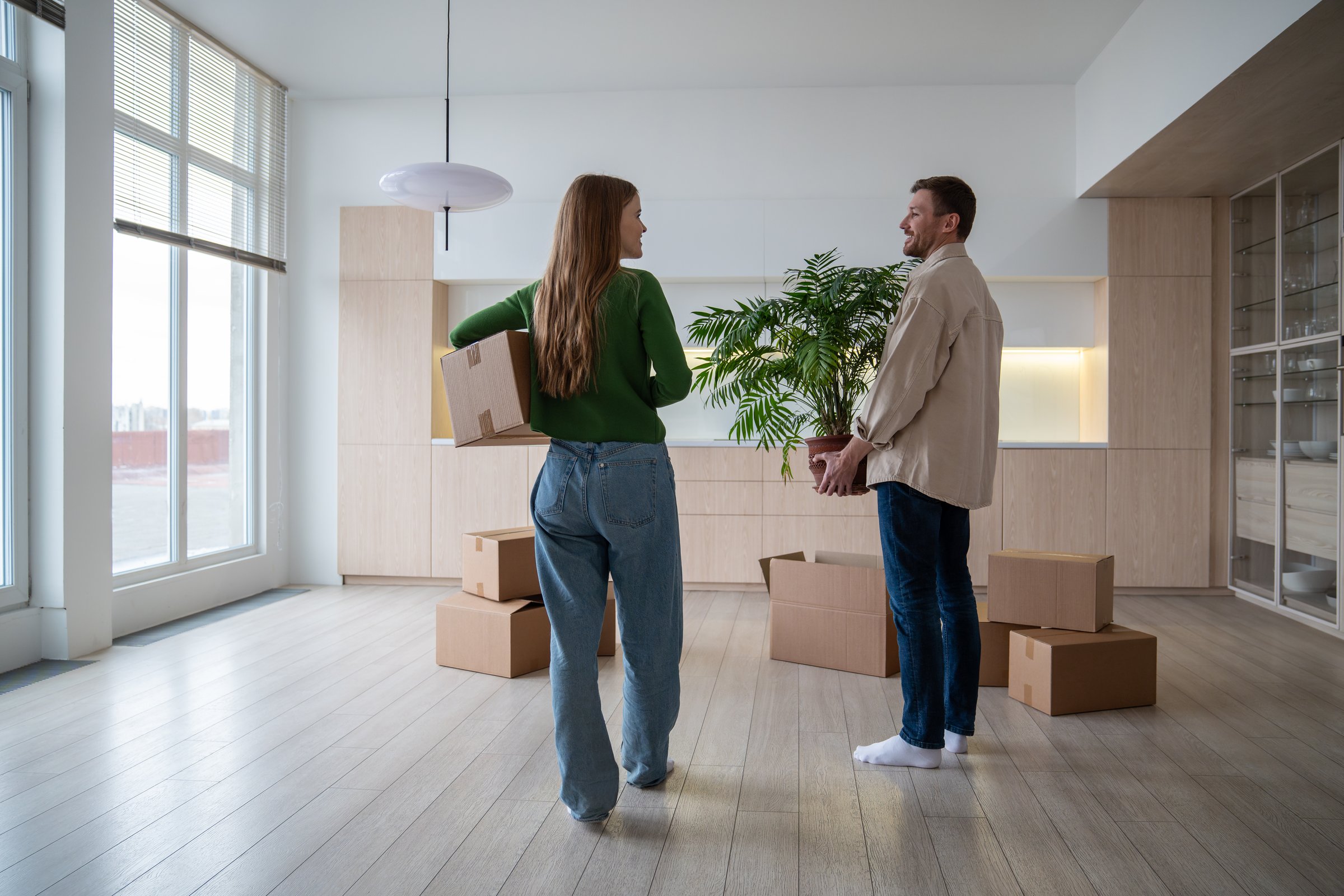 Happy joyful couple standing in spacious empty flat, speaking while resting after carrying houseplants, heavy cardboard boxes with personal things, belongings. Relocation day into own new apartment