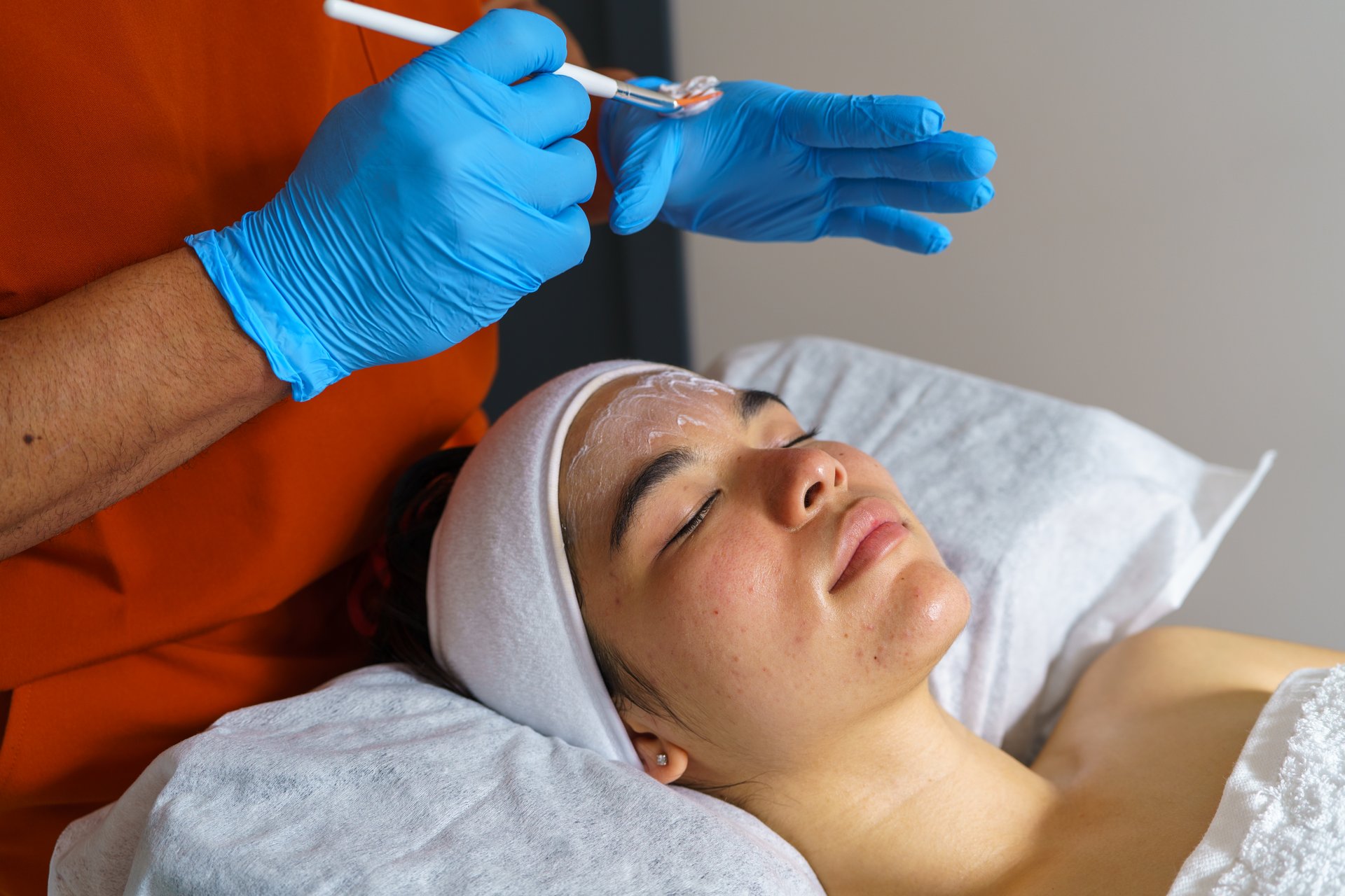 Beautician applying a facial mask with a brush on a young asian woman's forehead, lying on a bed, for an acne treatment