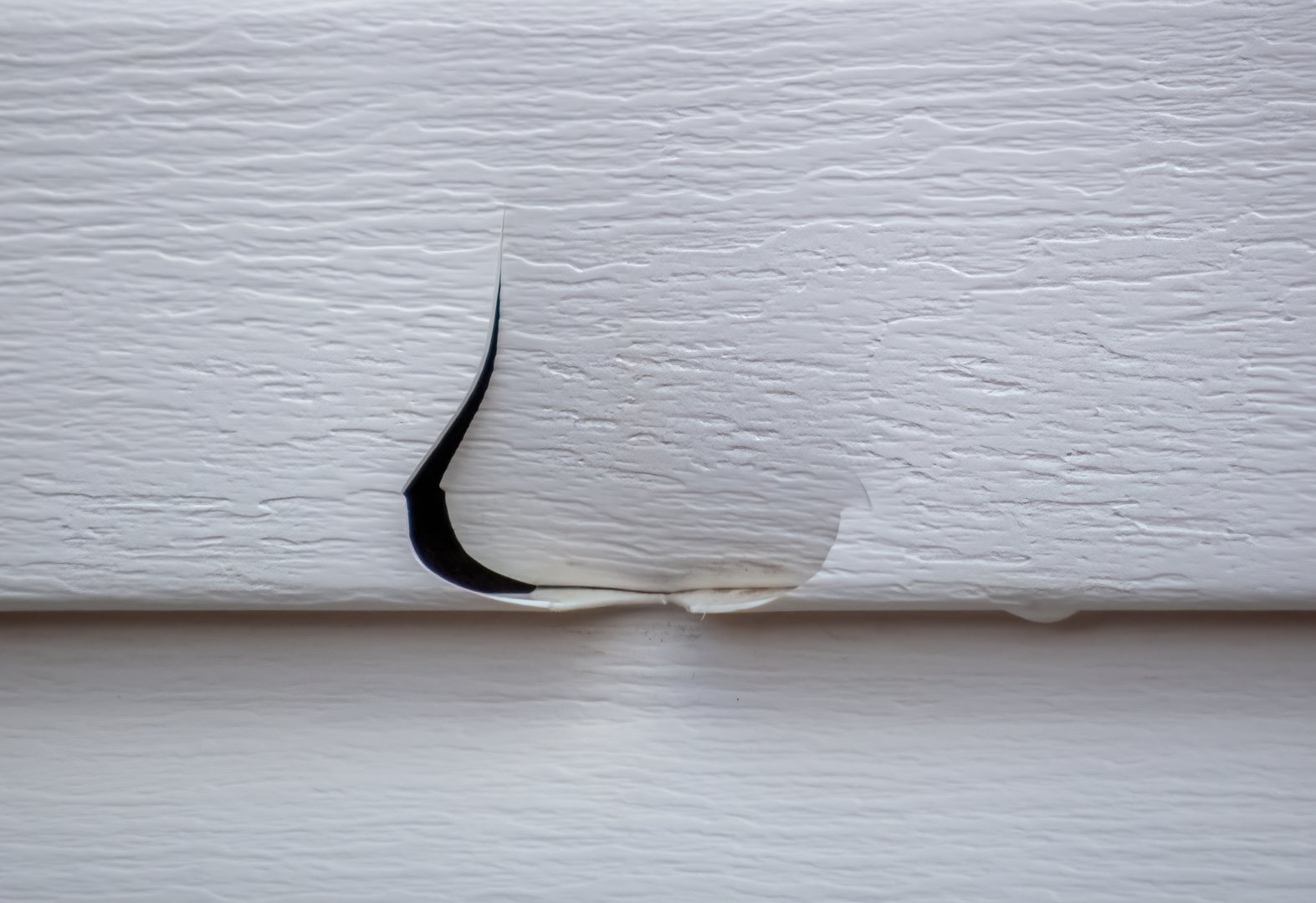A small, tear-shaped hole has appeared in a white vinyl siding panel, indicating potential hail damage or structural issues.