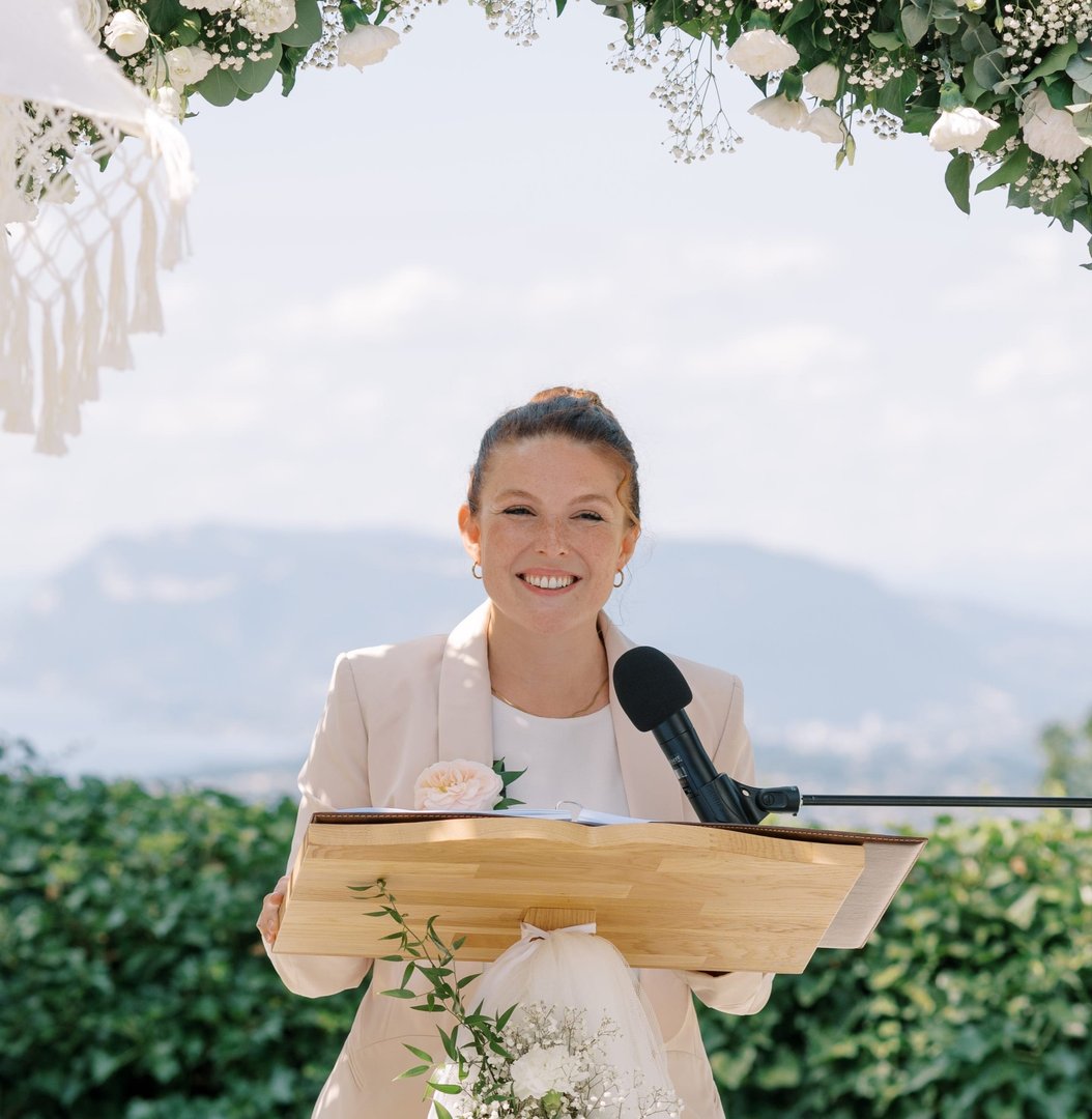 Woman smiles while speaking at a podium outdoors, surrounded by flowers and greenery, with mountains in the background.