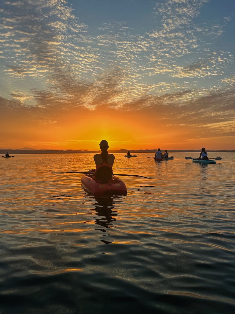Silhouettes of people kayaking on a calm lake at sunset, with a vibrant orange sky and scattered clouds.