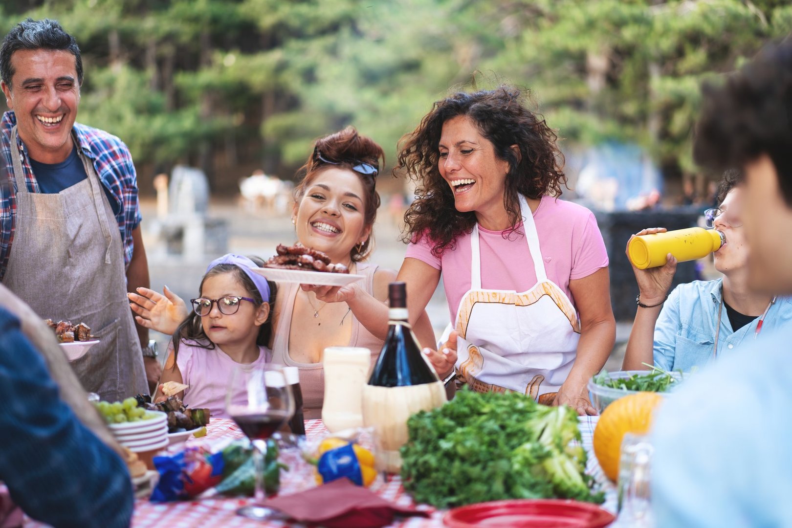 Candid shot of diverse family enjoying outdoor barbecue. Multigenerational group laughing and sharing food at picnic table in forest. Concept of family bonding, summer gatherings, and joyful moments.