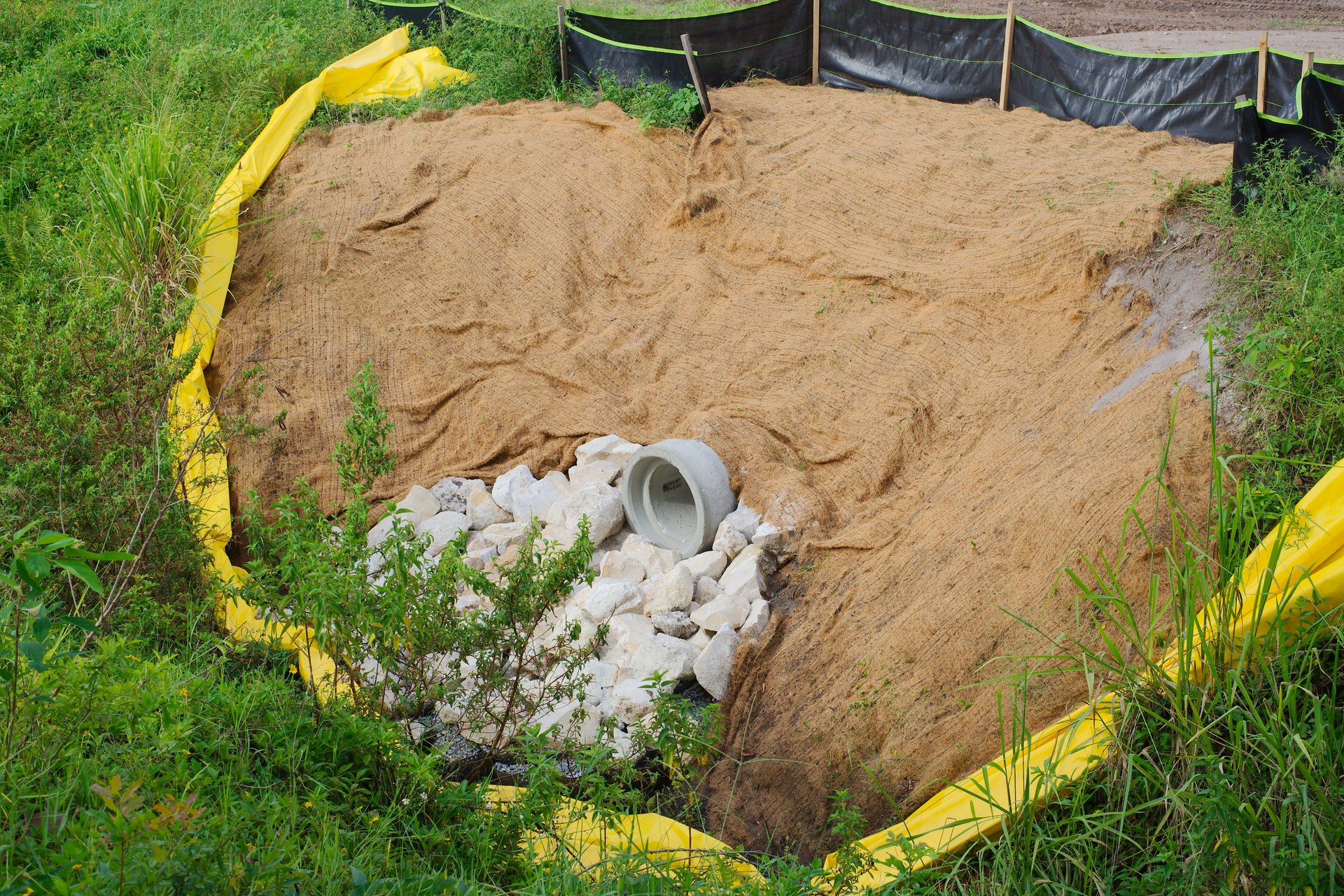 A sediment control basin featuring a pipe outlet, rocks, silt fencing, and erosion matting, set in a natural grassy environment, showcasing effective erosion prevention and water management techniques.