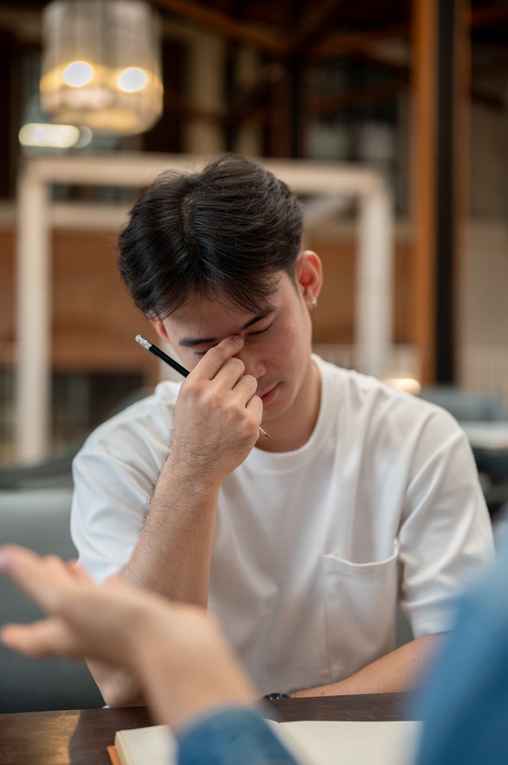 A serious, stressed Asian man is listening to his colleague discuss work-related problems as they work together to find a solution, expressing frustration and a headache.