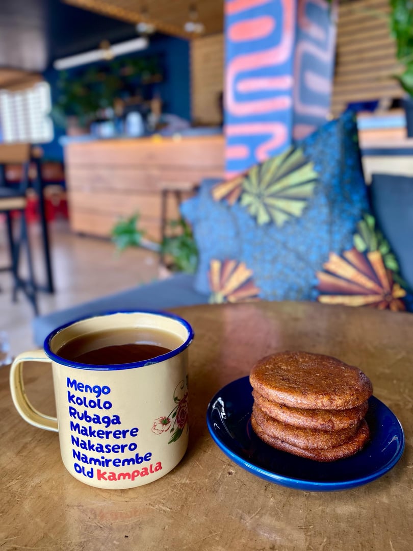 A mug with Kampala neighborhood names and four brown cookies on a blue plate on a wooden table in a cozy cafe setting.