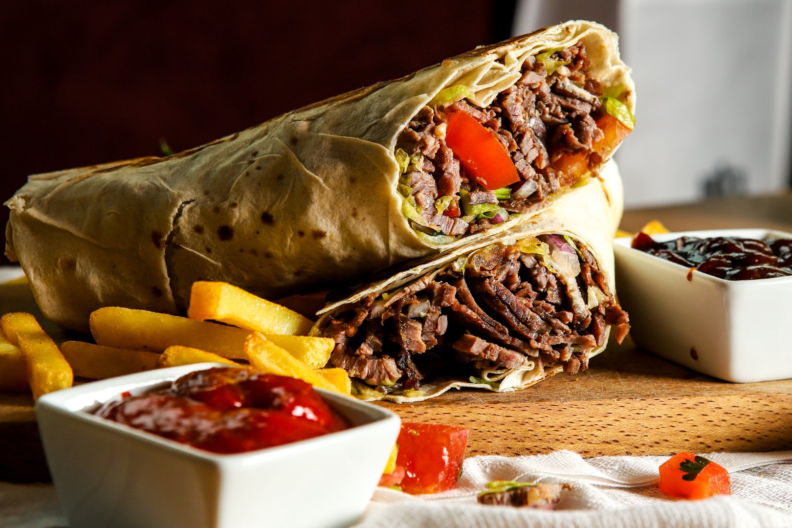 A delicious and hearty beef shawarma wrap, sliced in half to reveal a generous filling of tender, seasoned meat, fresh lettuce, and ripe tomatoes. The meal is served on a rustic wooden board, accompanied by a side of crispy, golden french fries and two small white bowls of dipping sauces, likely ketchup and barbecue sauce. The close-up shot emphasizes the textures and savory appeal of this popular and flavorful Middle Eastern street food dish.
