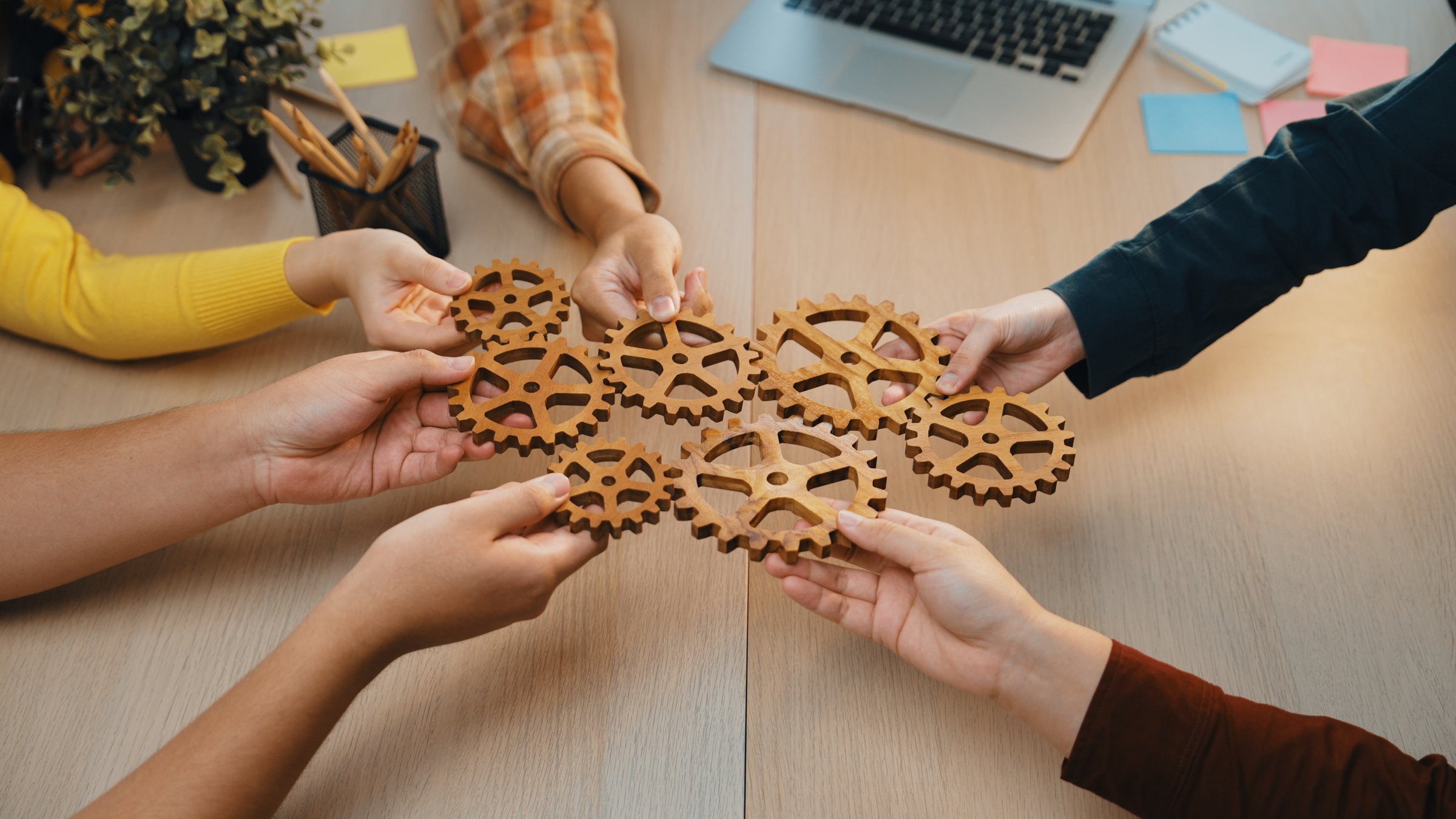 A group of diverse hands holds wooden gears in a collaborative workspace, symbolizing teamwork and innovation during a planning session in a modern office. SACTR