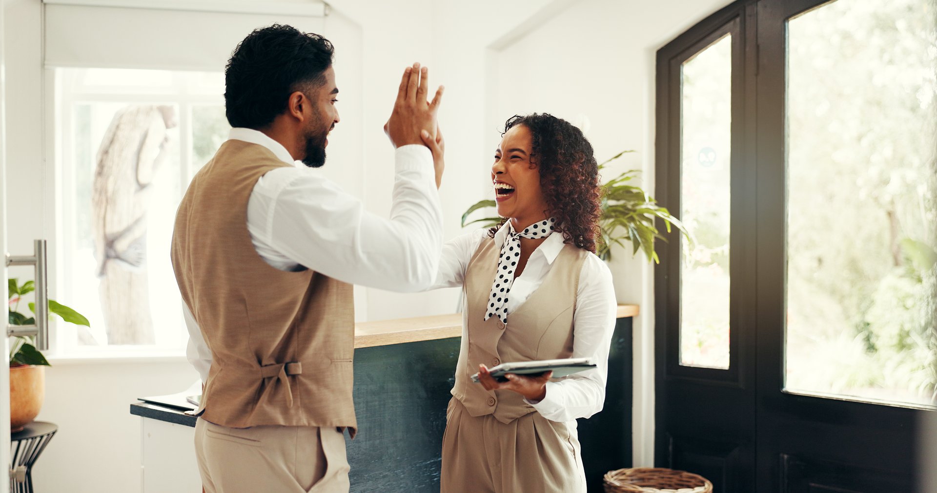 High five, online booking and tablet with hotel concierge staff in lobby together for celebration. App, hospitality and success with reception team in lodge or resort for accommodation planning