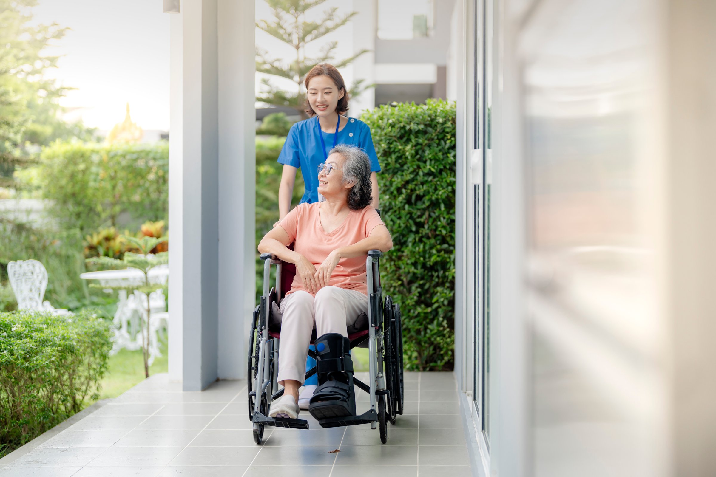 An elderly woman in a wheelchair is assisted by a healthcare professional outdoors. The scene emphasizes care, support, and companionship in a peaceful, green environment.