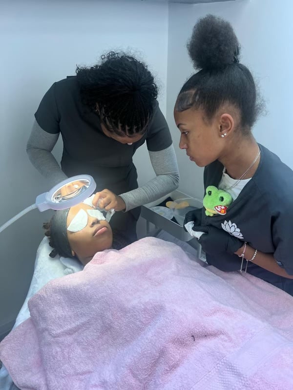 A person receiving skincare treatment on a bed, covered with a pink towel, assisted by two people, one holding a plush toy.