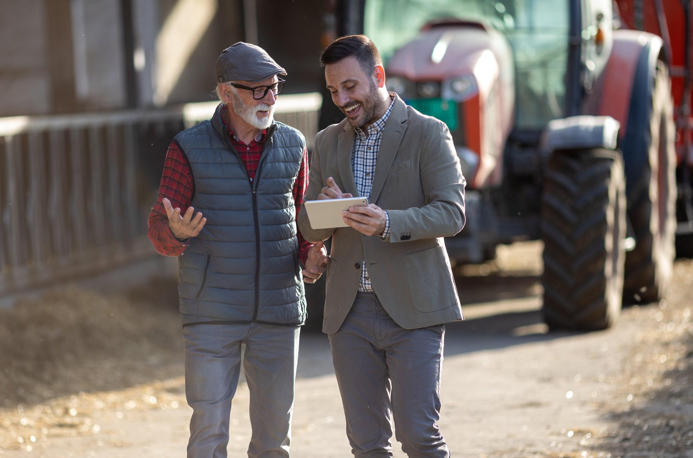 Bank agent and senior farmer talking about business and looking at tablet in front of tractor on cattle ranch