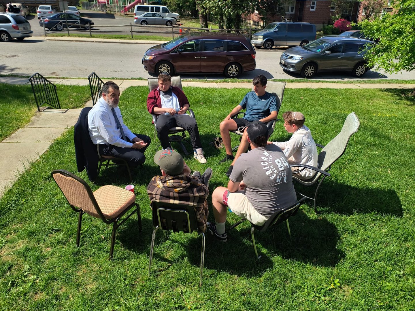 A group of six men sit in a circle of chairs on a lawn, having a discussion on a sunny day.