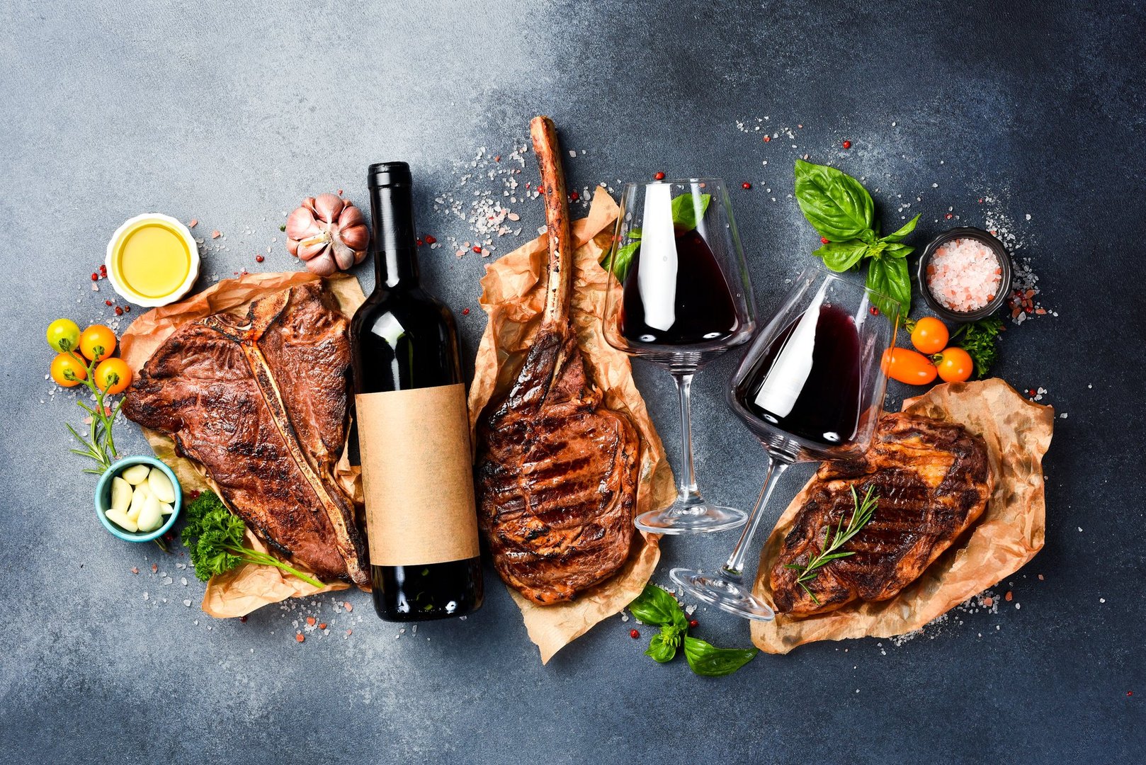 Glasses of red wine and a set of Black Angus beef steaks, on a gray stone background. Top view, close up. Banner. Free space for text.