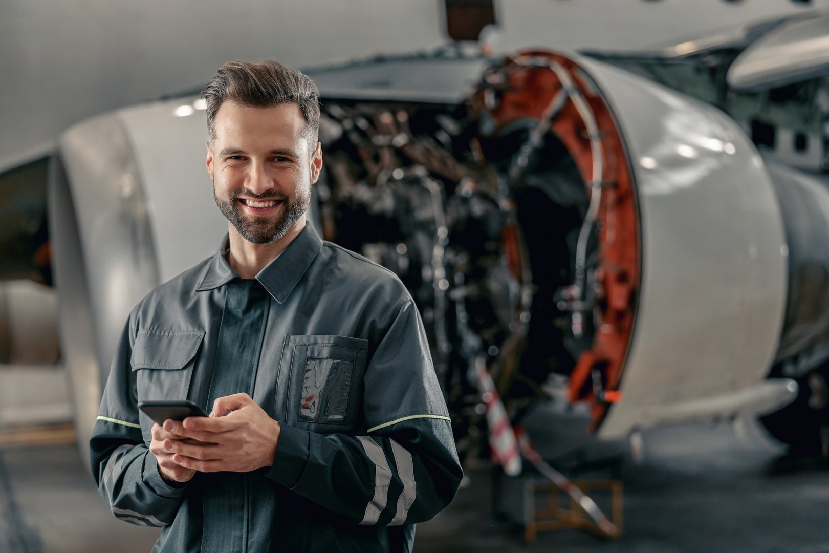 Joyful bearded man aircraft maintenance technician looking at camera and smiling while holding smartphone