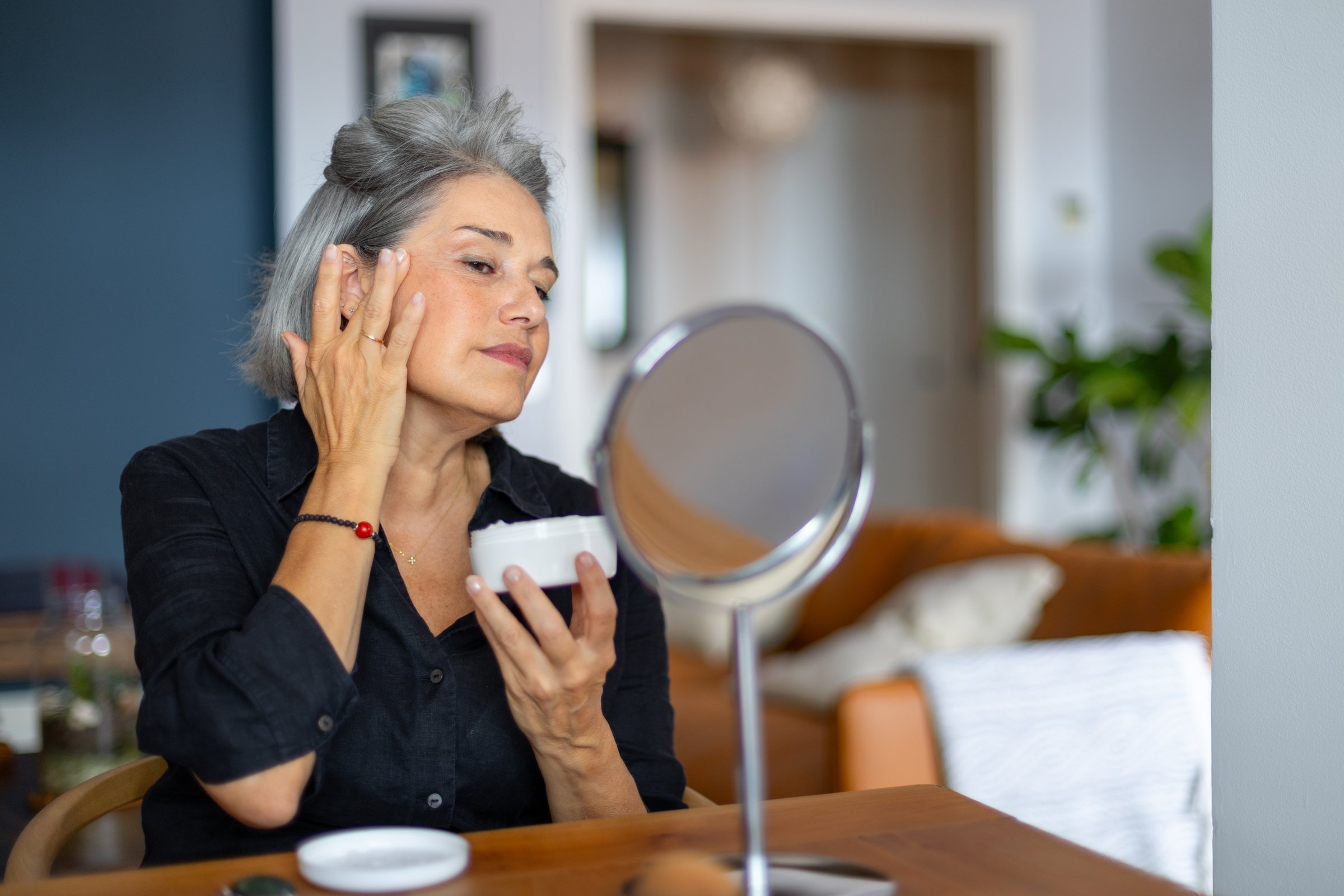 Mature woman taking care of her skin, applying anti aging cream while looking in a small mirror