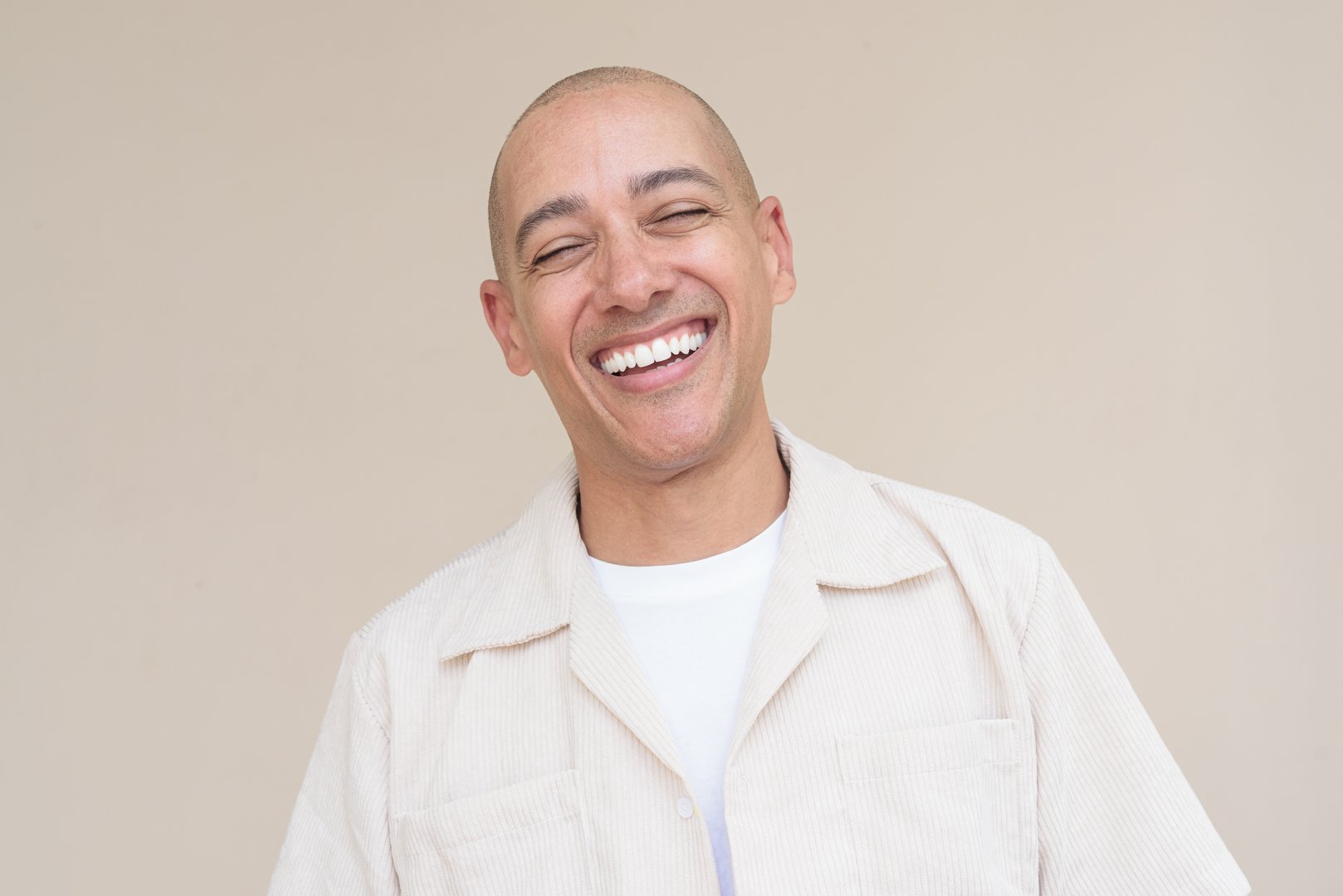 Portrait of bald Hispanic man wearing casual beige shirt standing against plain beige background.