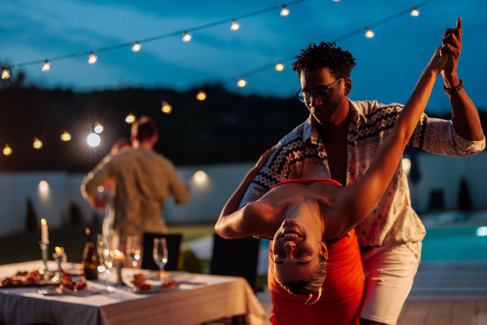 Young couple dancing at a night party near swimming pool with string lights and friends in the background
