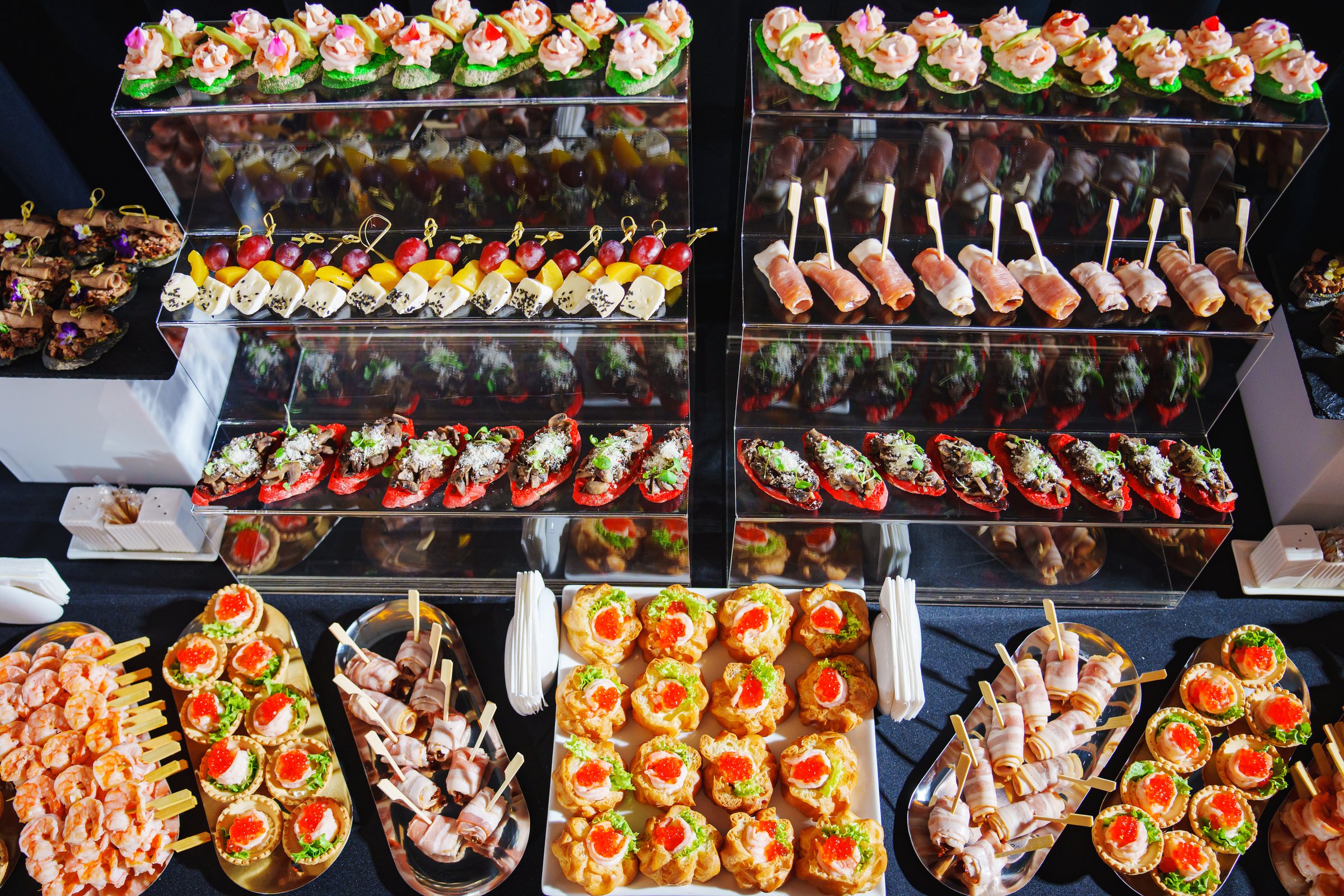 A colorful display of appetizers showcases a variety of culinary delights at a large gathering. The table features finger foods such as shrimp, skewers, and assorted canapеs, enticing guests
