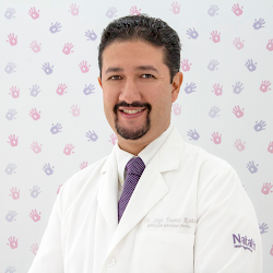A smiling doctor in a white coat and purple tie stands in front of a wall with colorful handprints.