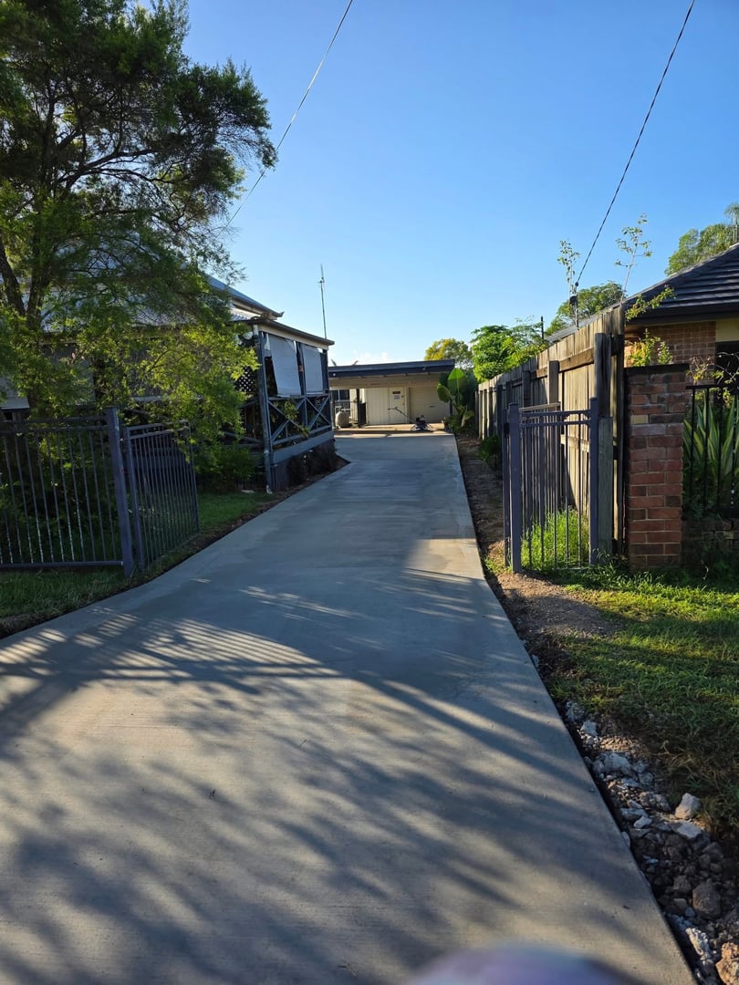 Long concrete driveway flanked by trees and fences, leading to a garage with clear blue sky above.