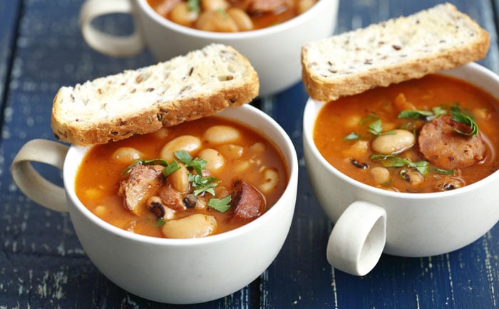 Two white cups of bean soup with sausage, garnished with herbs, topped with multigrain bread, on a rustic blue table.