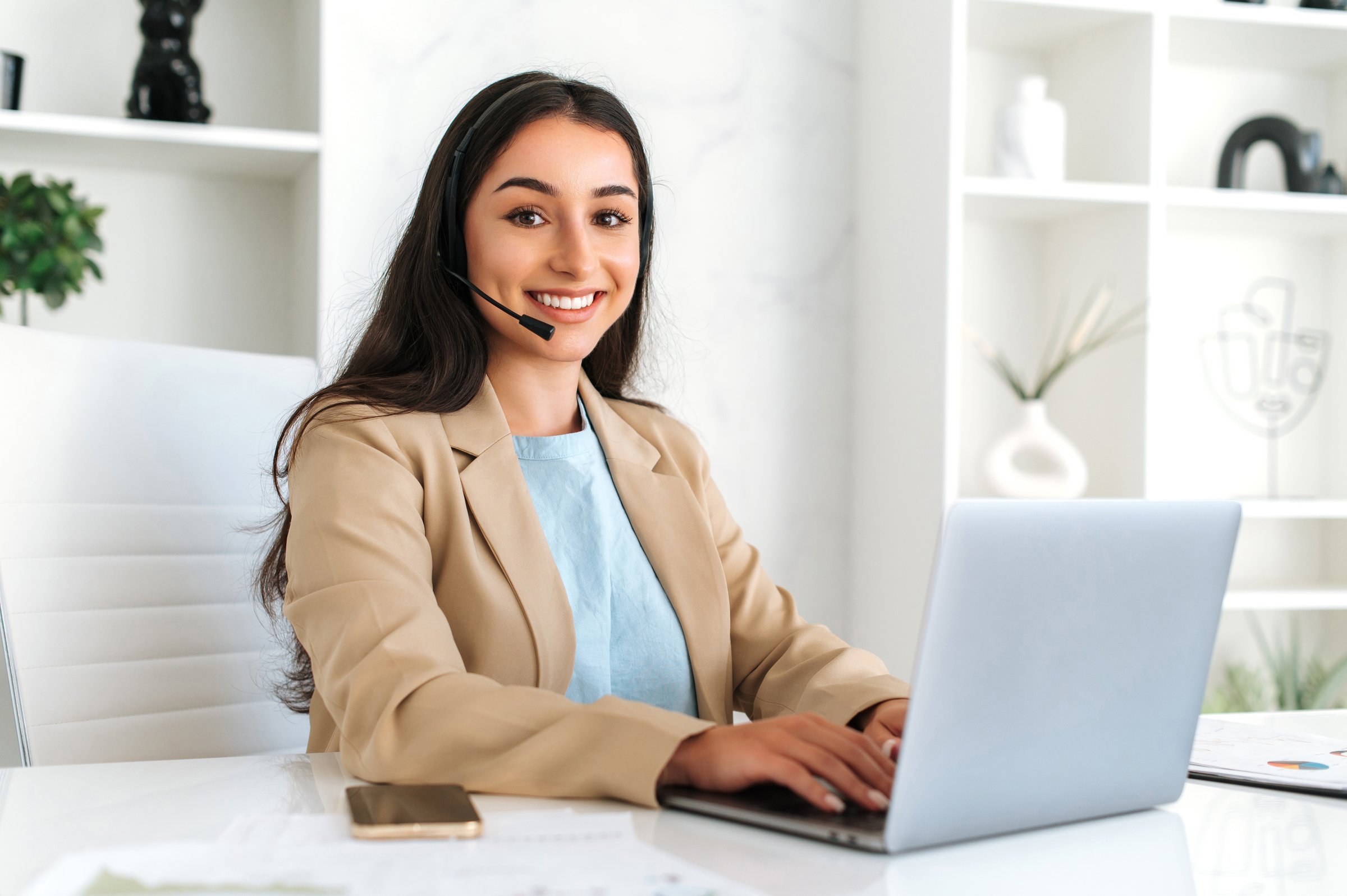 Call center, customer support service. Confident positive woman, call center operator with headset, working on support hotline in a modern office with a laptop, looking at the camera, smiling friendly