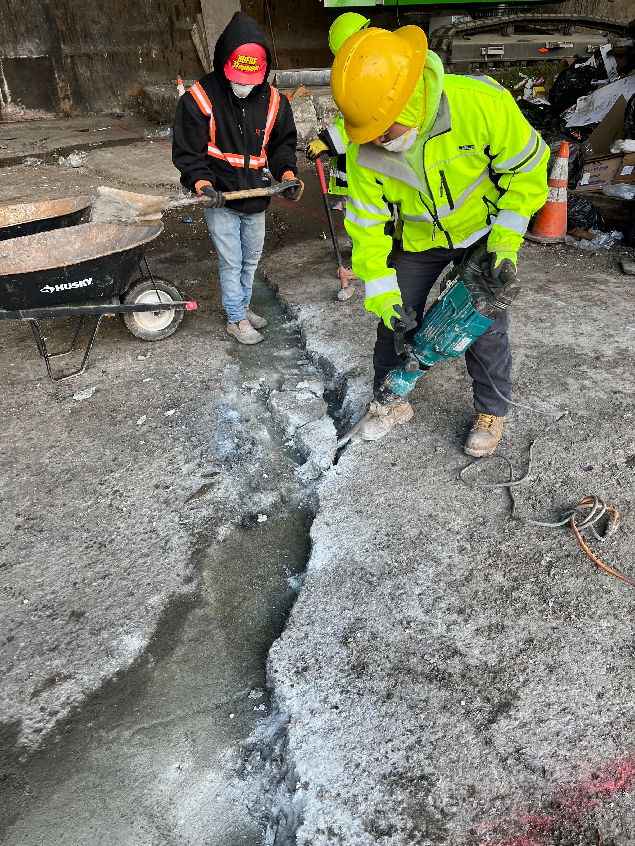 Two workers in safety gear using a jackhammer to break concrete in a construction site, surrounded by debris and tools.