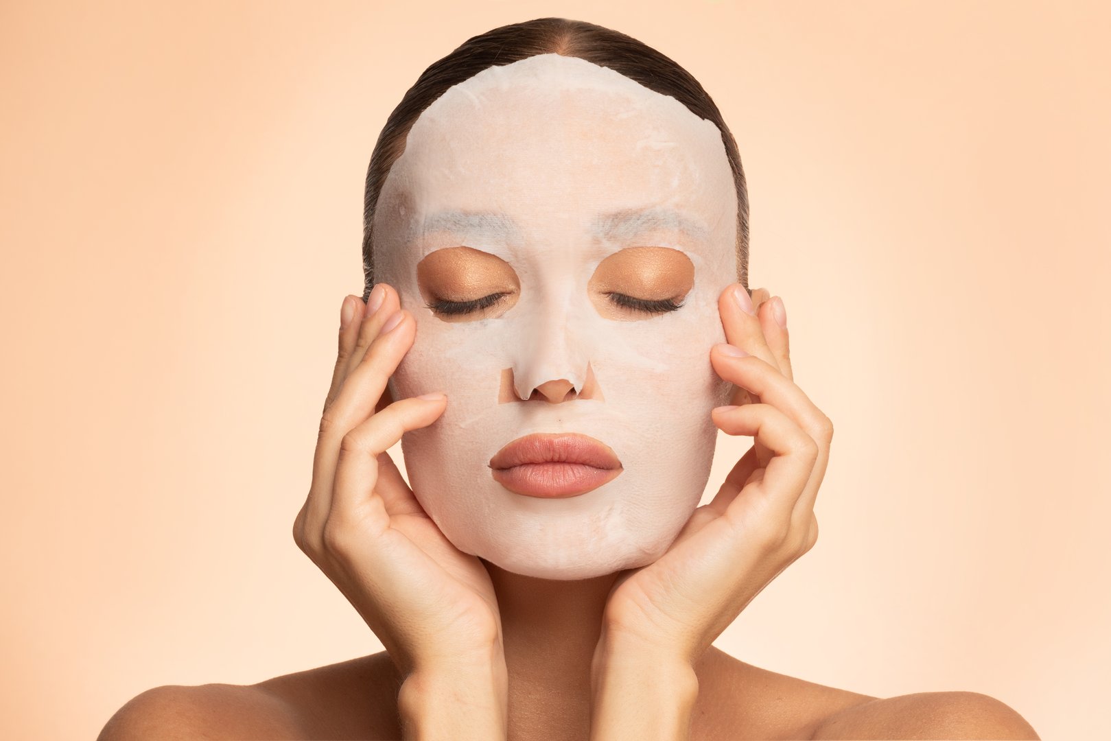 Young European woman with eyes closed, applying cotton facial moisturizing mask on face, takes care of skin, posing over beige background