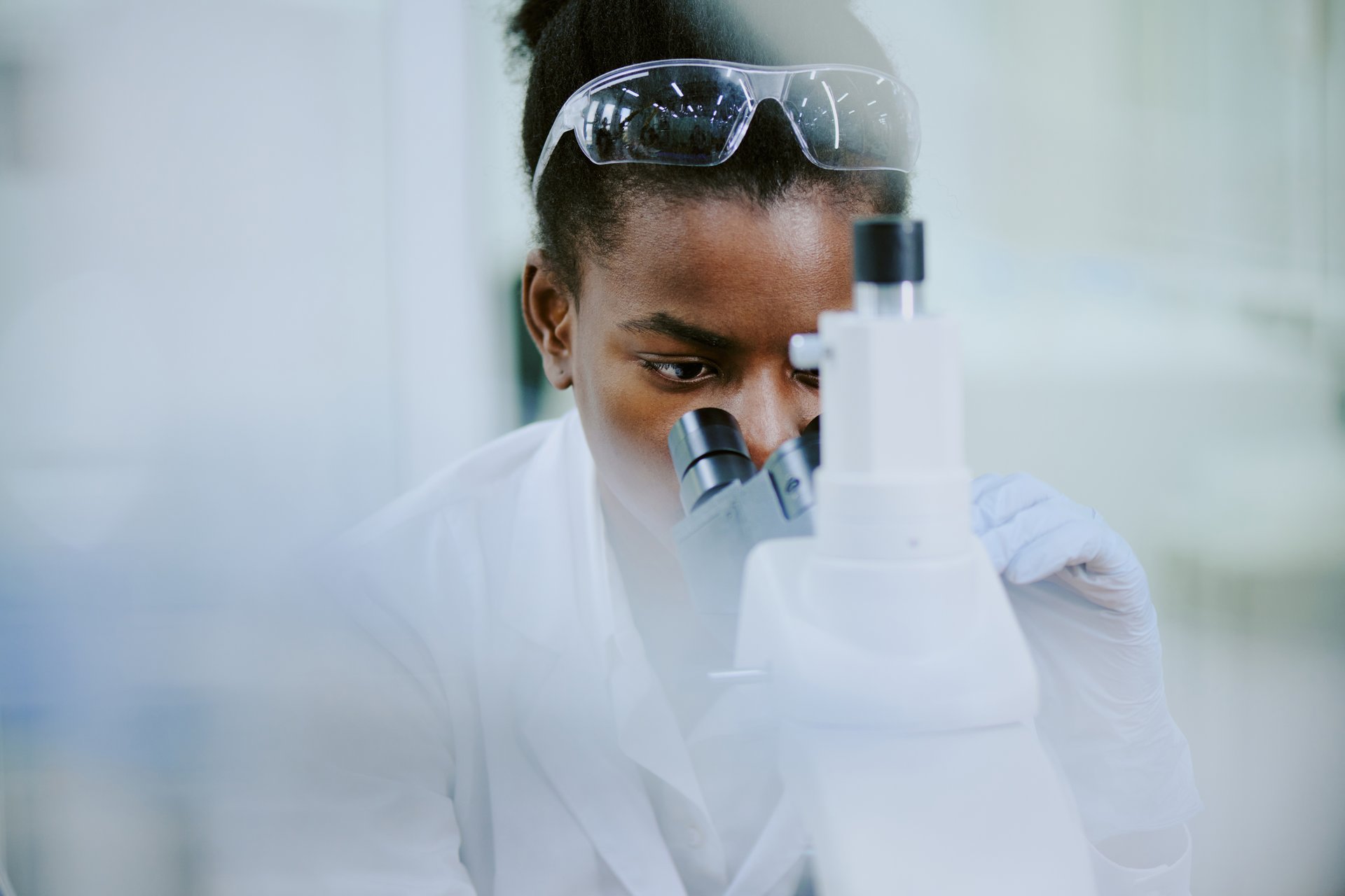 African American female scientist working in laboratory focusing on microscopic examination wearing lab coat and safety glasses carefully analyzing results with precise concentration