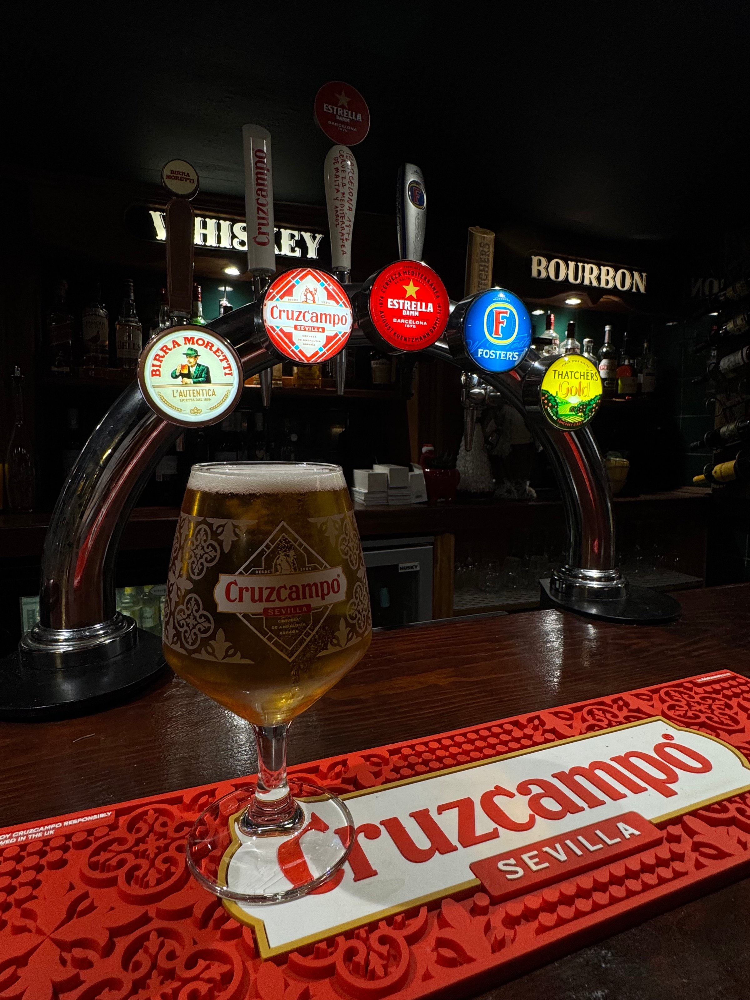 Cruzcampo beer glass on bar counter with illuminated beer taps behind in a dimly lit bar.