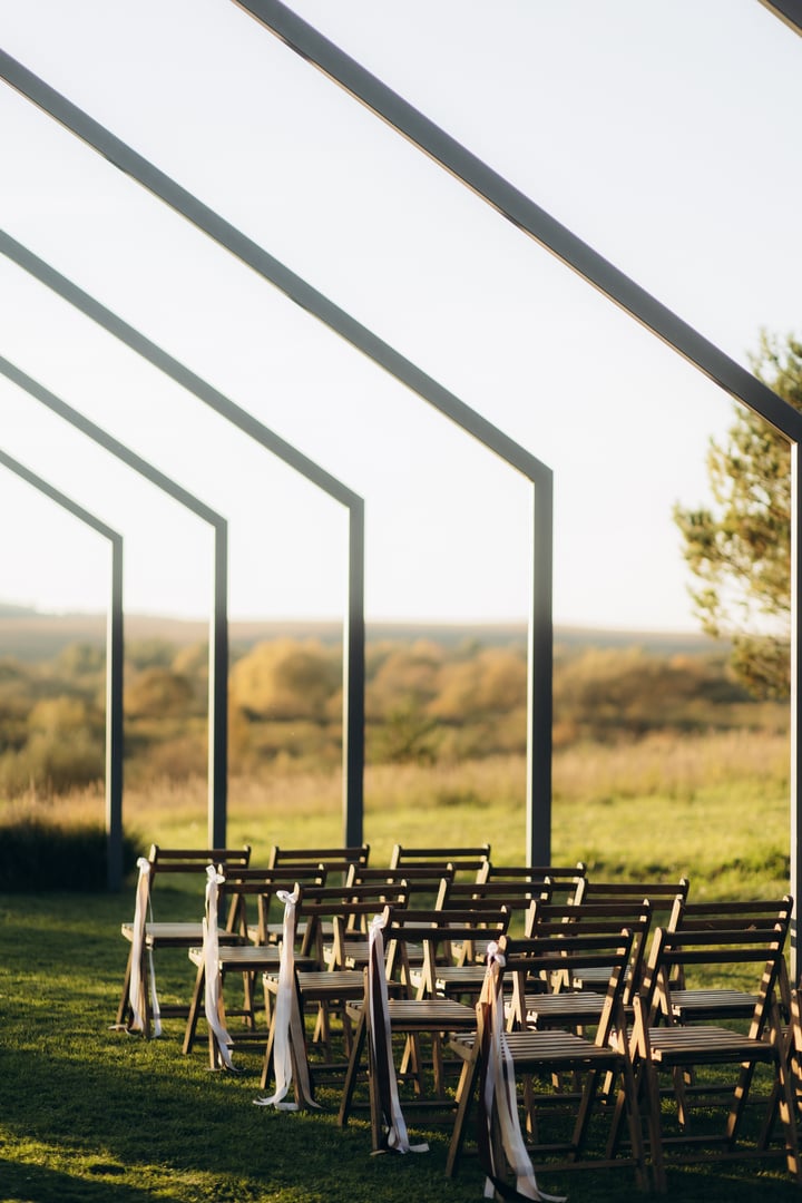 Beautiful outdoor venue for a wedding ceremony featuring rows of wooden chairs adorned with white ribbons, placed in a scenic, natural, open setting with golden-hour lighting and a serene environment.