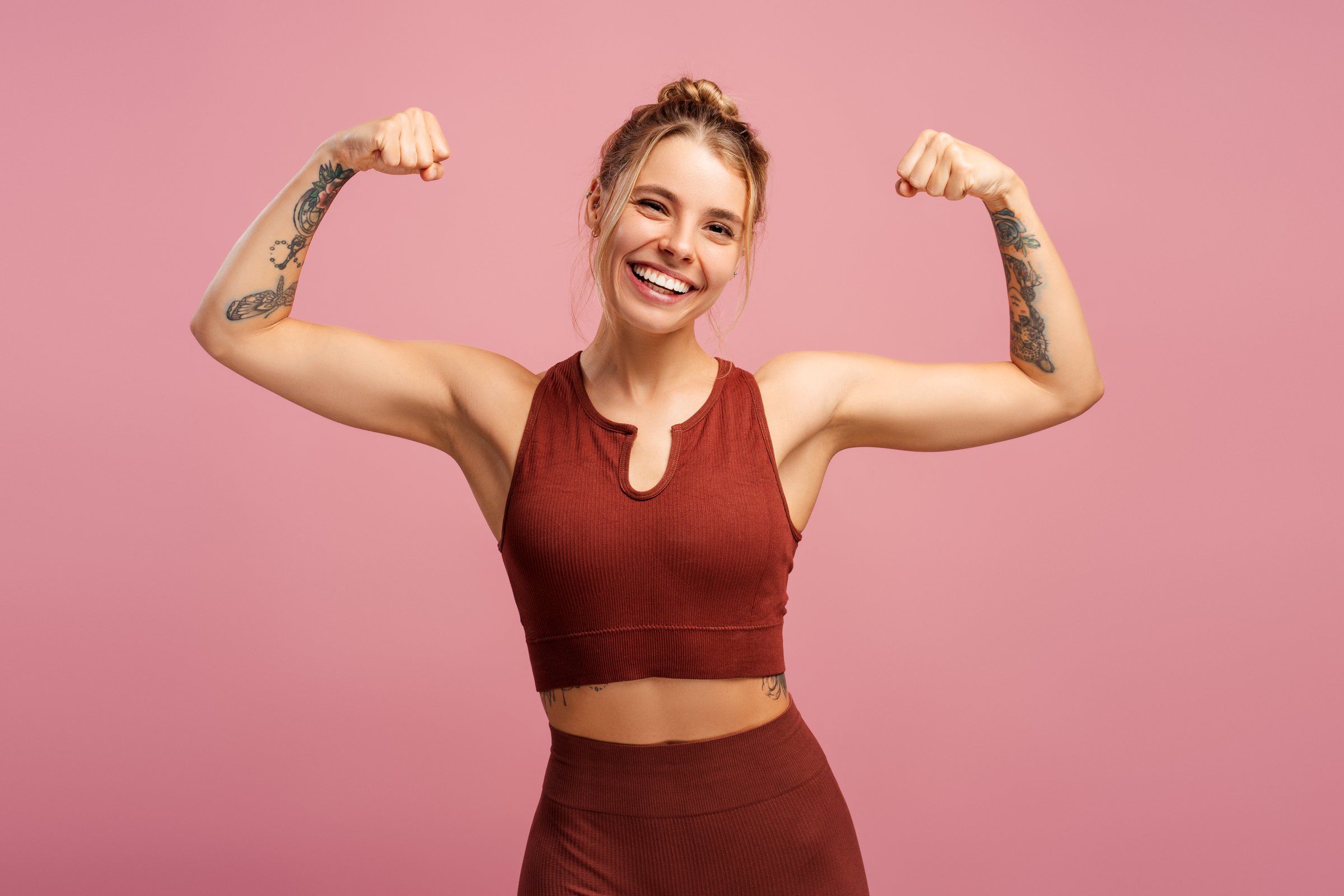 Portrait of positive sporty young woman showing muscles on her arms, looking at camera, isolated on pink. Sport, health care, wellness concept