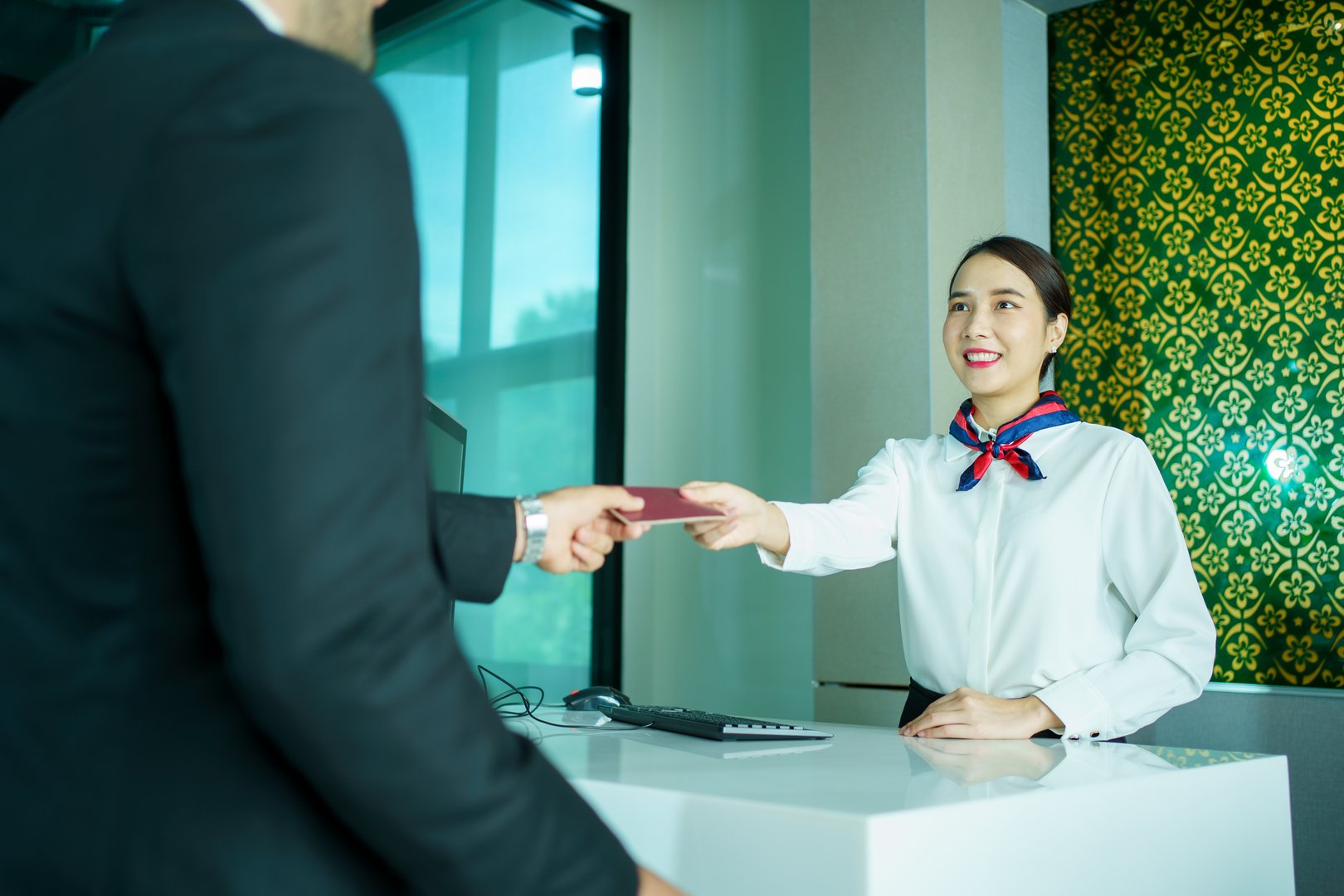 An Asian businessman hands over his passport and documents to a friendly airline check-in agent or hotel receptionist, engaging in polite and professional conversation during his travel check-in process.
