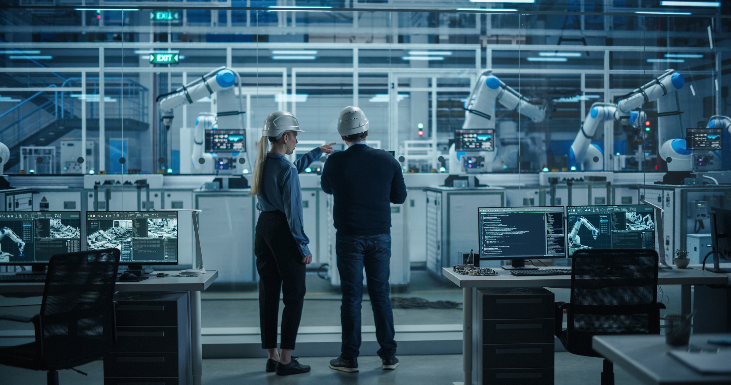 Engineer and Technician Standing with Their Back to Camera, Having a Discussion and Using a Laptop Computer. Industrial Specialists Analyzing Conditions at an Electronics Factory with an Assembly Line