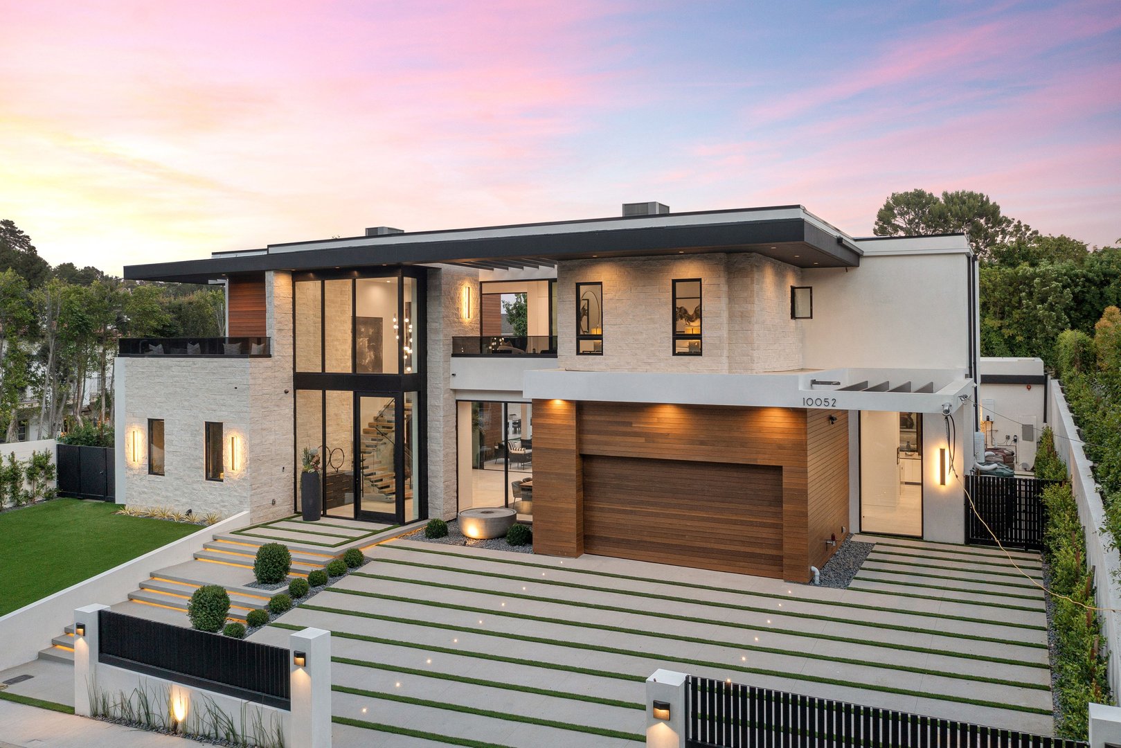 Modern luxury home exterior with large windows, wooden garage door, and well-manicured lawn at sunset.