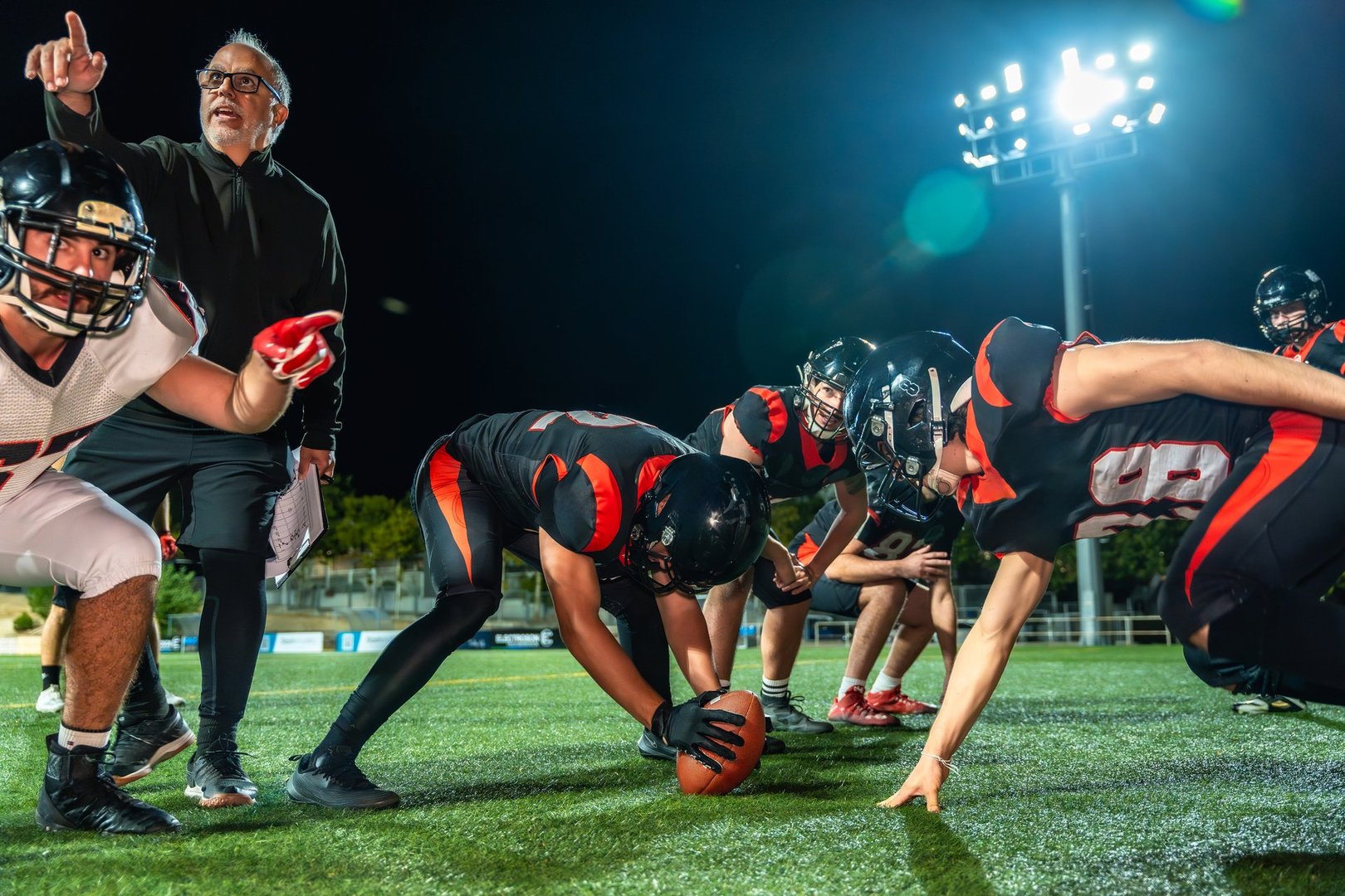 American football coach instructing players on the field during a night game, with one player holding the ball at the line of scrimmage, preparing for action